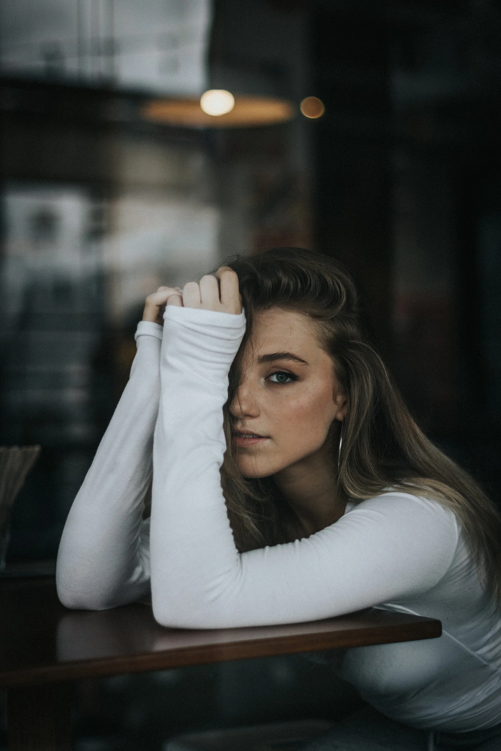 A woman with long brown hair and blue eyes sitting at a dark wood table in a dimly lit modern cafe, resting her head on her left arm, wearing a white long-sleeve shirt, with her right hand gently touching her forehead.