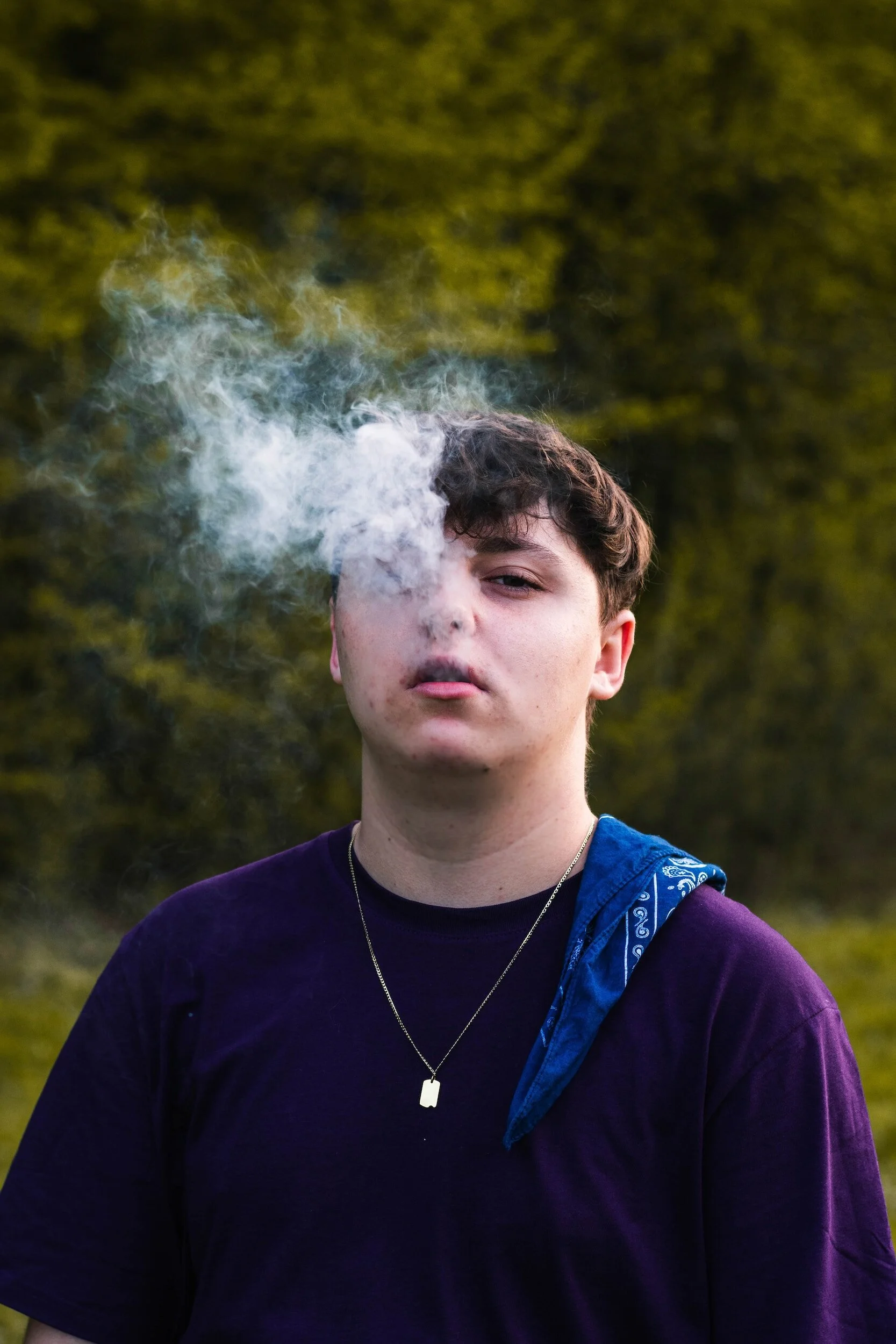 A young person with short, dark hair standing outdoors, exhaling smoke with a blurred natural background of trees and greenery.