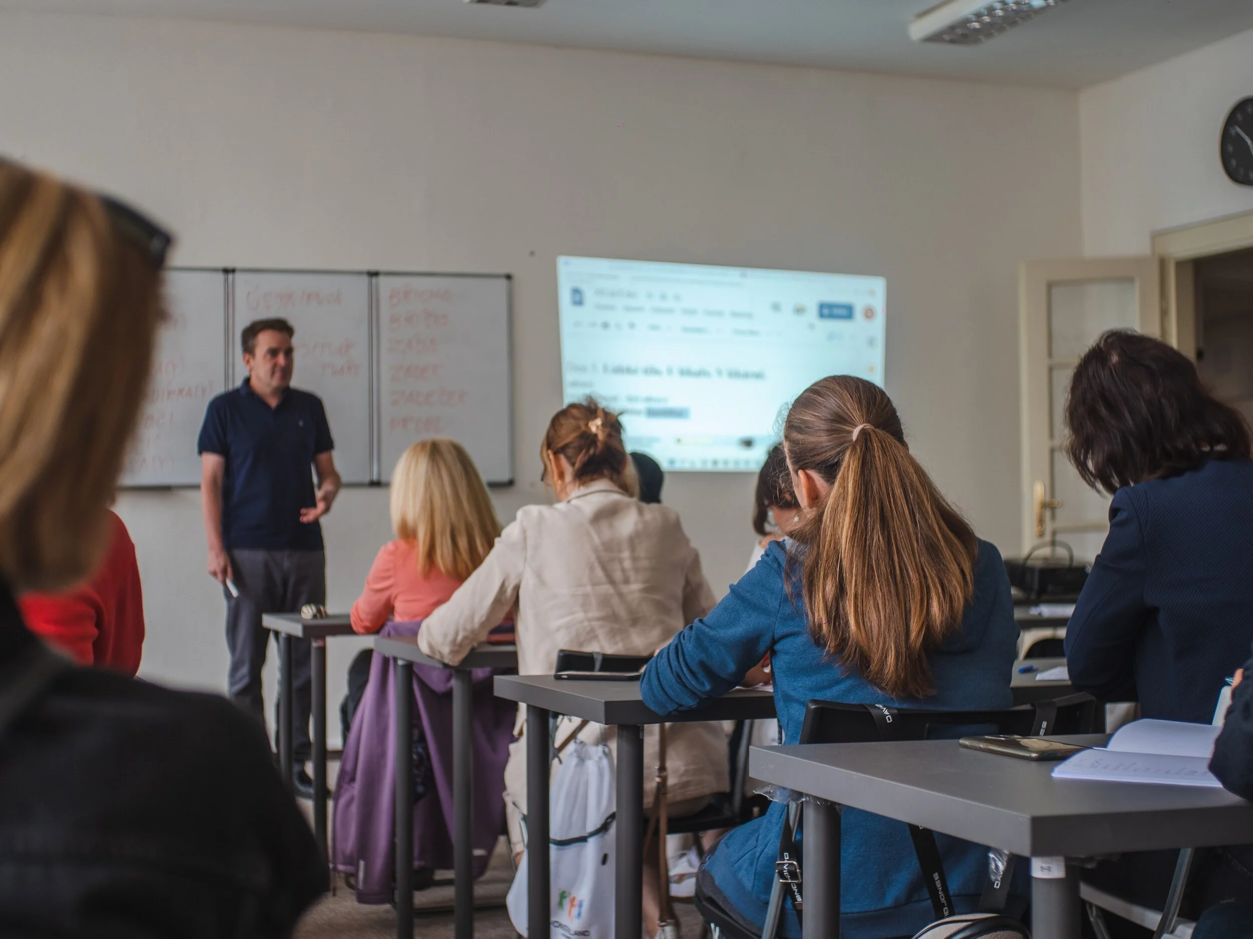A classroom with a teacher standing at the front near a whiteboard and a projection screen, while students sit at desks listening to the lesson.