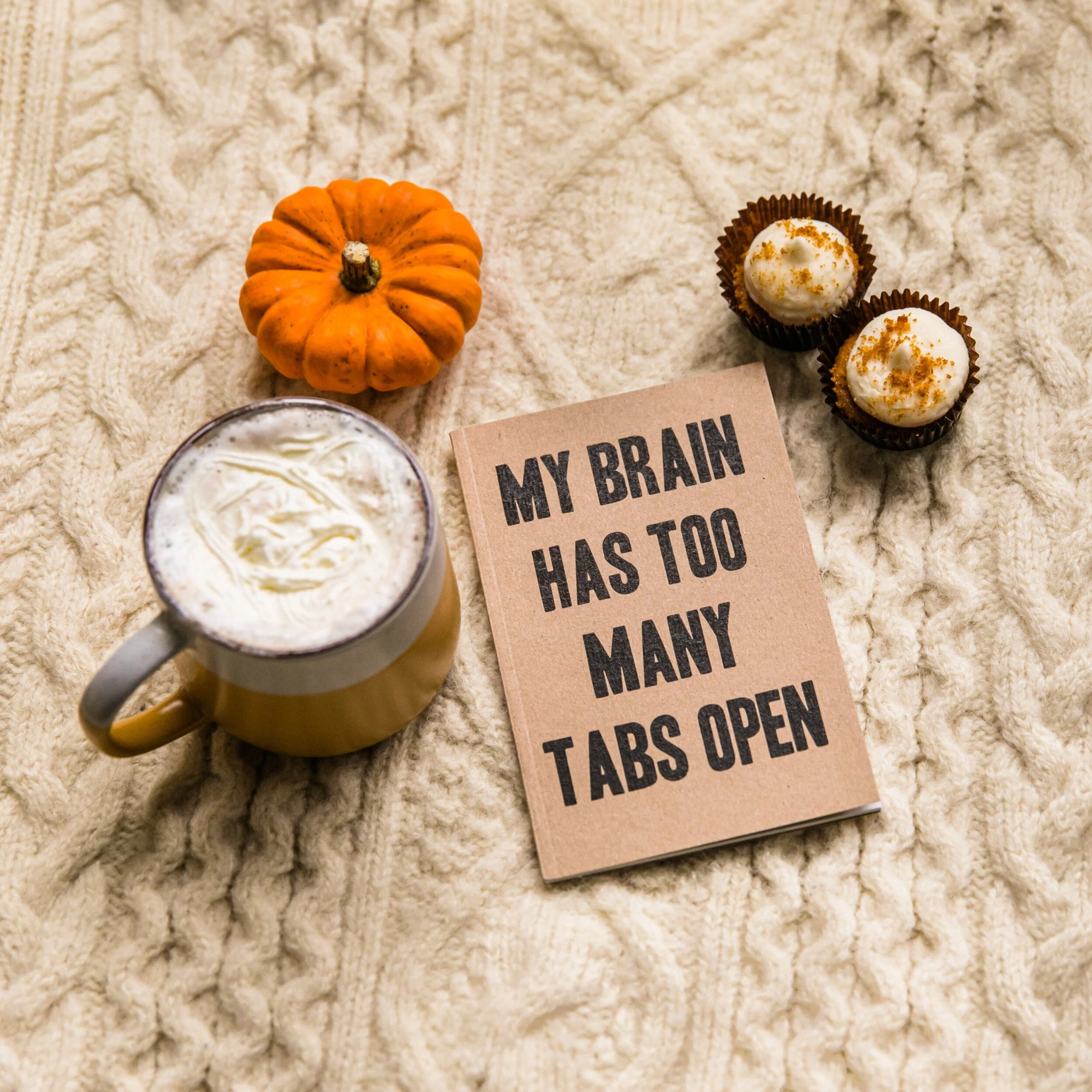 A cozy arrangement of a small pumpkin, two cupcakes with cream topping, a mug of whipped cream or whipped coffee, and a sign that reads 'MY BRAIN HAS TOO MANY TABS OPEN' on a knitted beige blanket.