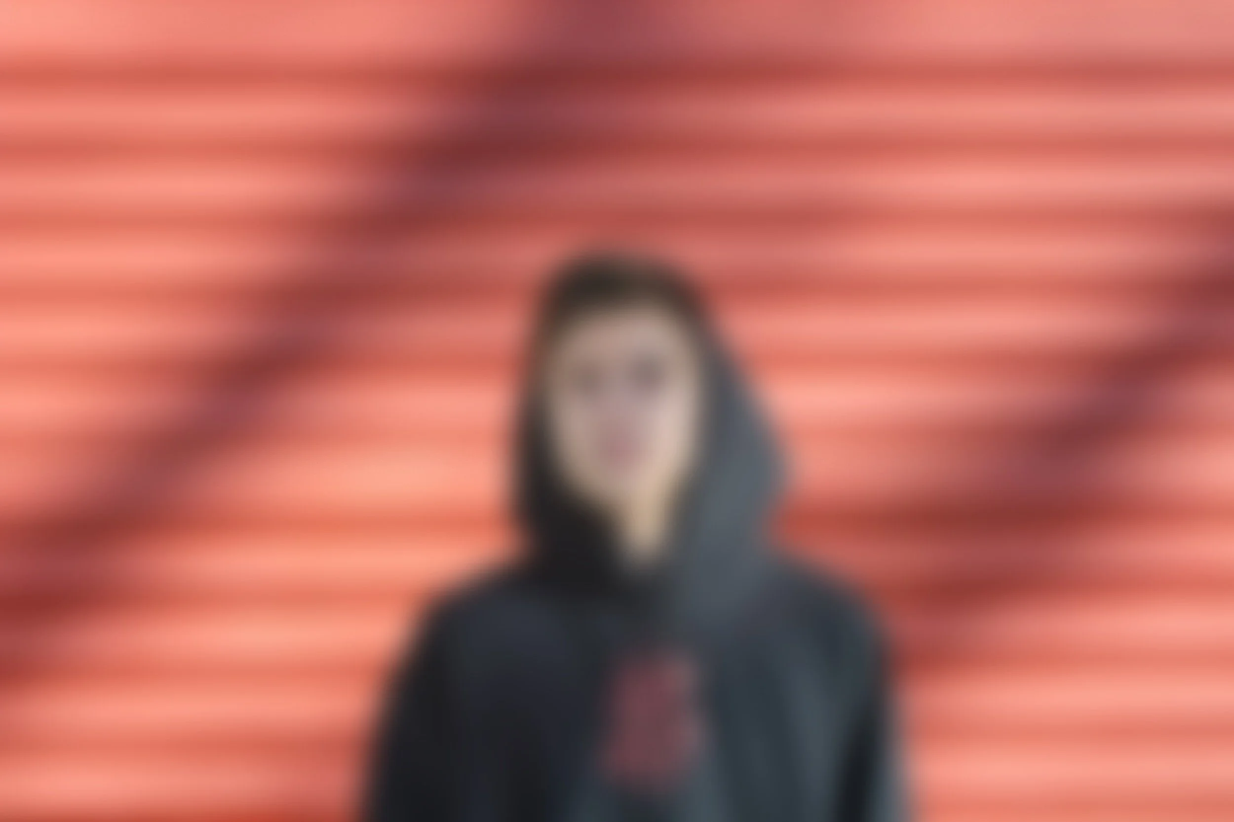 A person in a black hoodie standing in front of a background with horizontal red and black stripes, intentionally blurred.