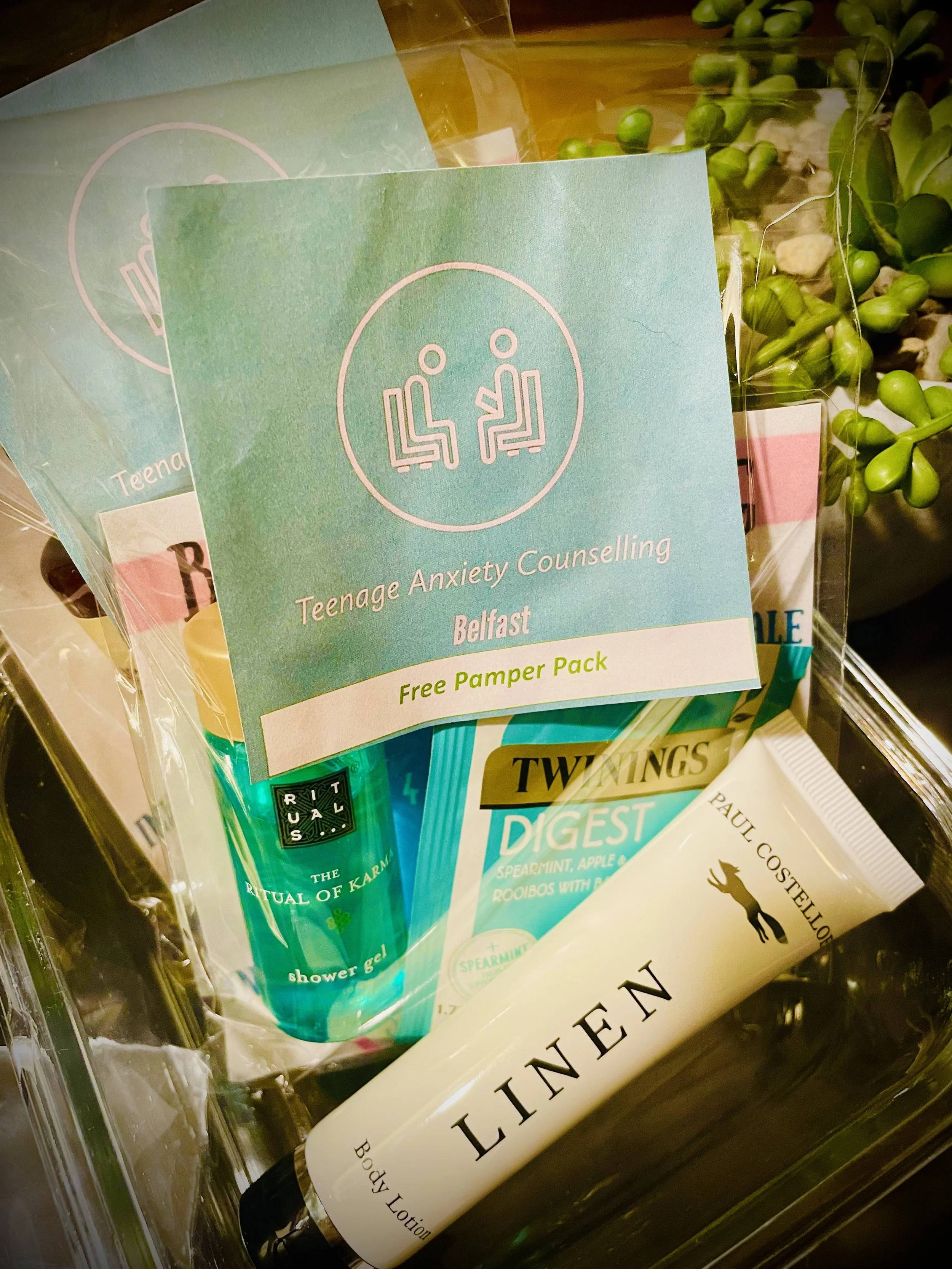 A gift basket containing a Teen Anxiety Counselling pamphlet, Twinings Digest herbal tea, and Linnen body lotion, with some potted plants in the background.
