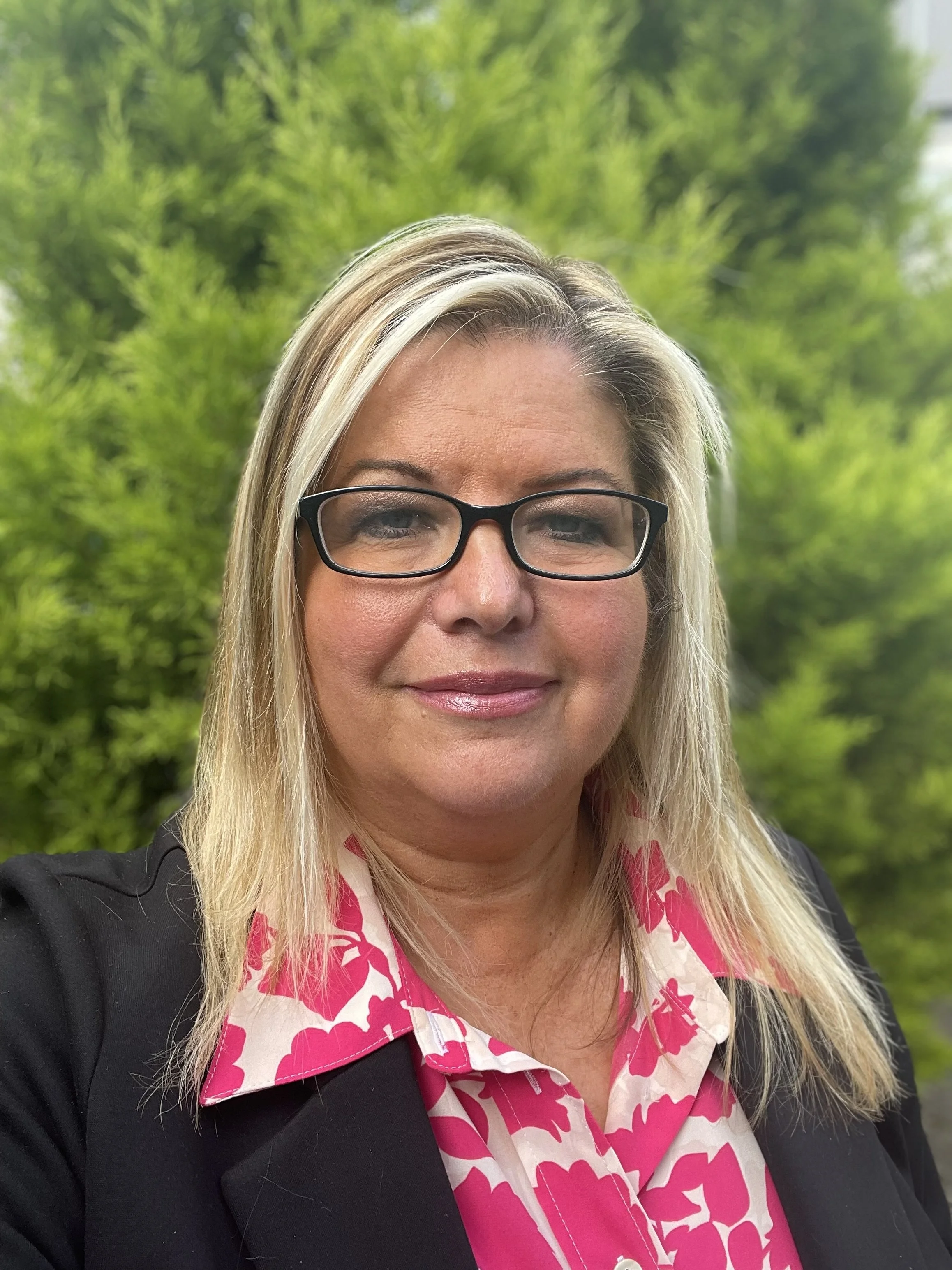 Close-up selfie of a smiling woman with blonde hair, wearing glasses, a black blazer, and a pink floral shirt, standing outdoors with green trees in the background.