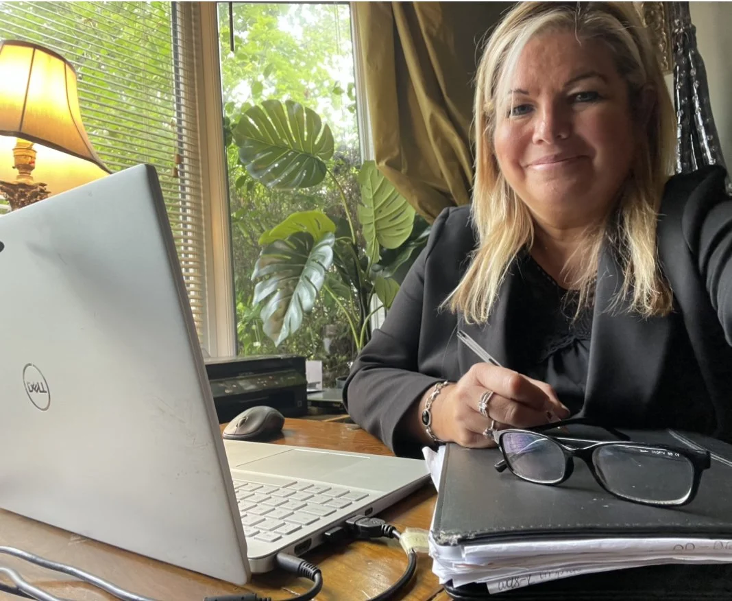 A woman with blonde hair sitting at a desk in a well-lit room, smiling at the camera, with a laptop, glasses, notebooks, and a pen in her hand, near a window with greenery outside.