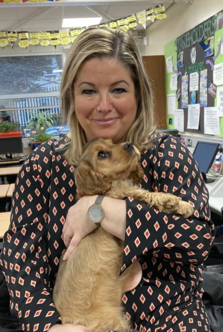 A woman with blonde hair wearing a patterned blouse holding a small brown dog in a classroom setting.