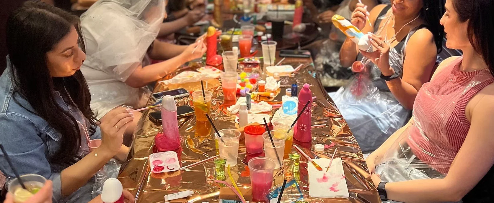 Group of women seated at a long table decorated with tape, engaging in a painting activity, with art supplies, drinks, and decorative items scattered on the table.