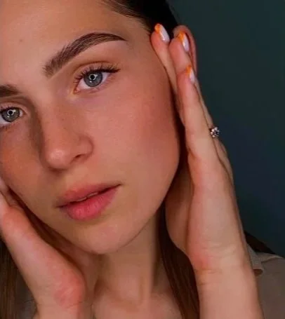 Close-up of a young woman with blue eyes and light makeup, holding her face with her hands, wearing a ring on her finger.