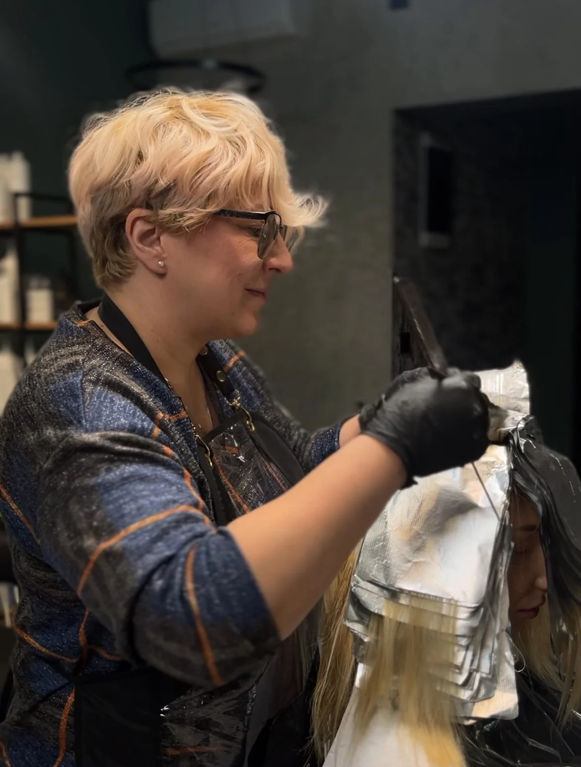 A woman with short blonde hair, glasses, and earrings is coloring a wig with black foil on a cap.