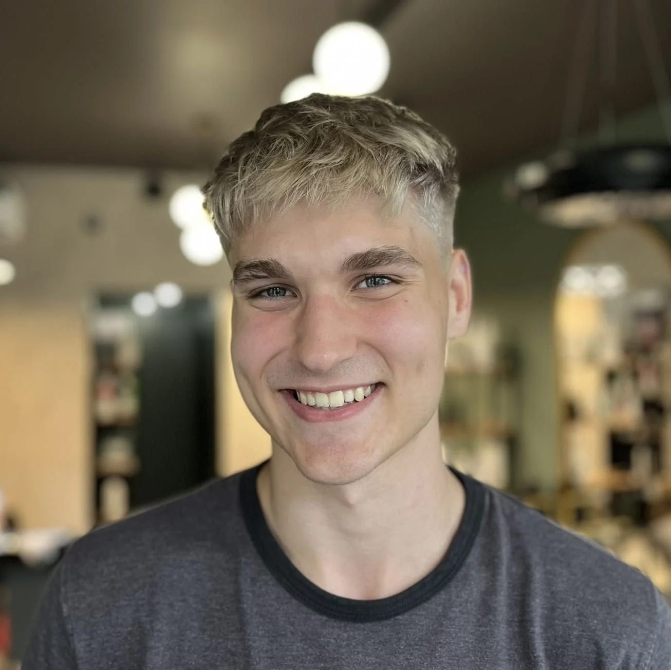 Smiling young man with blonde hair and blue eyes in a casual dark gray T-shirt in a cozy, well-lit indoor setting.