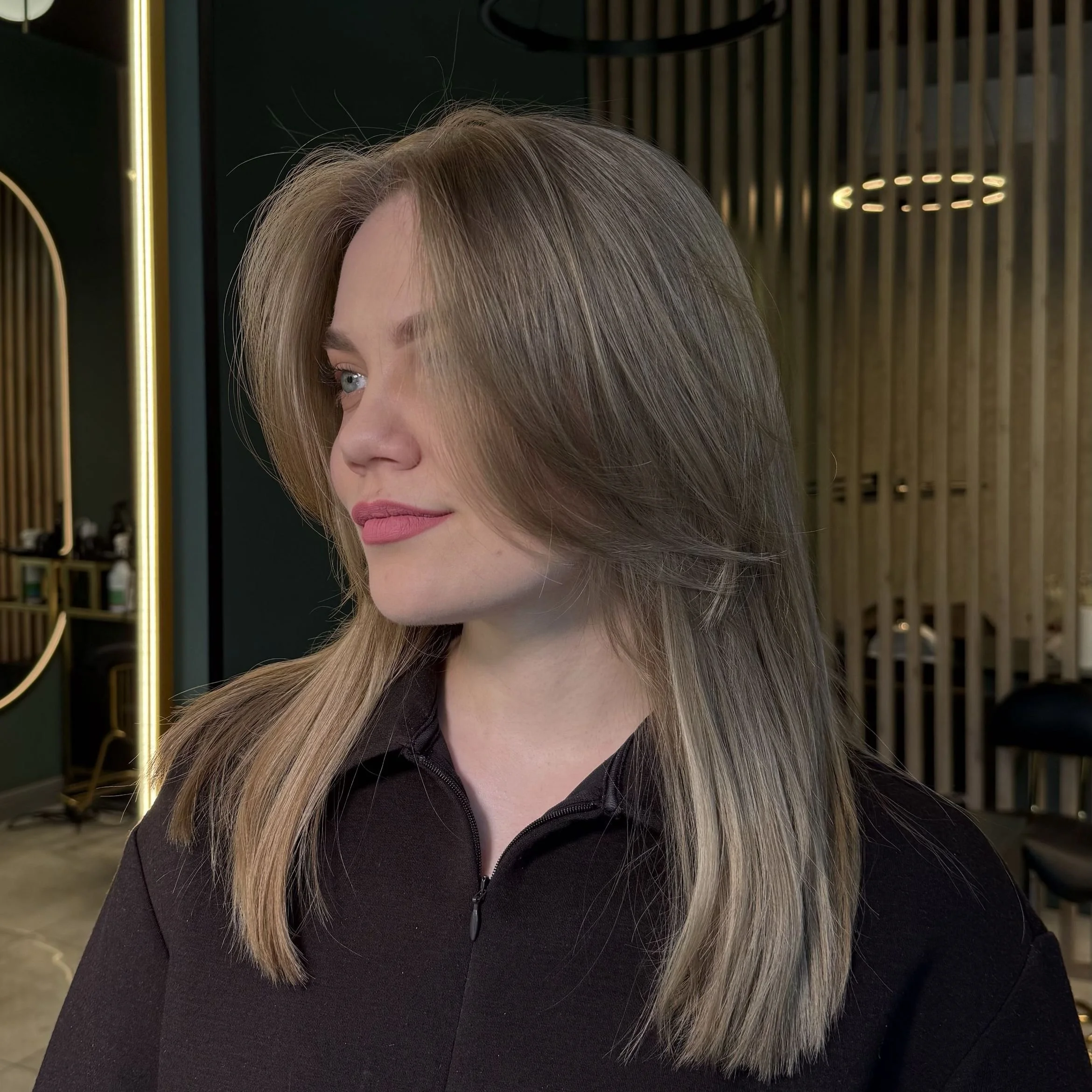 Woman with long, layered blonde hair, wearing a black zip-up top, in a modern salon interior.