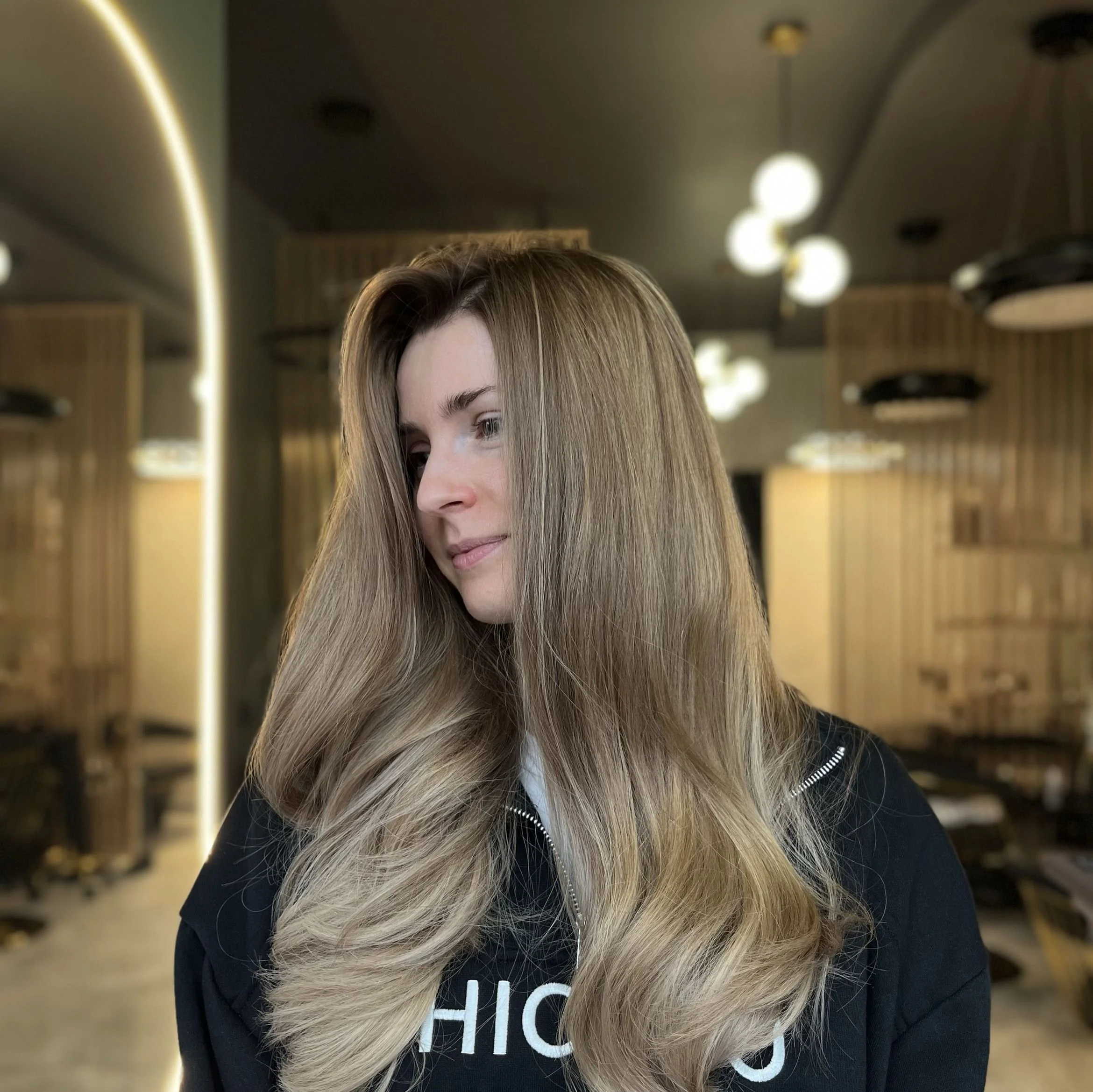 A woman with long, blonde, wavy hair standing in an indoor setting with modern decor and multiple spherical pendant lights hanging from the ceiling.