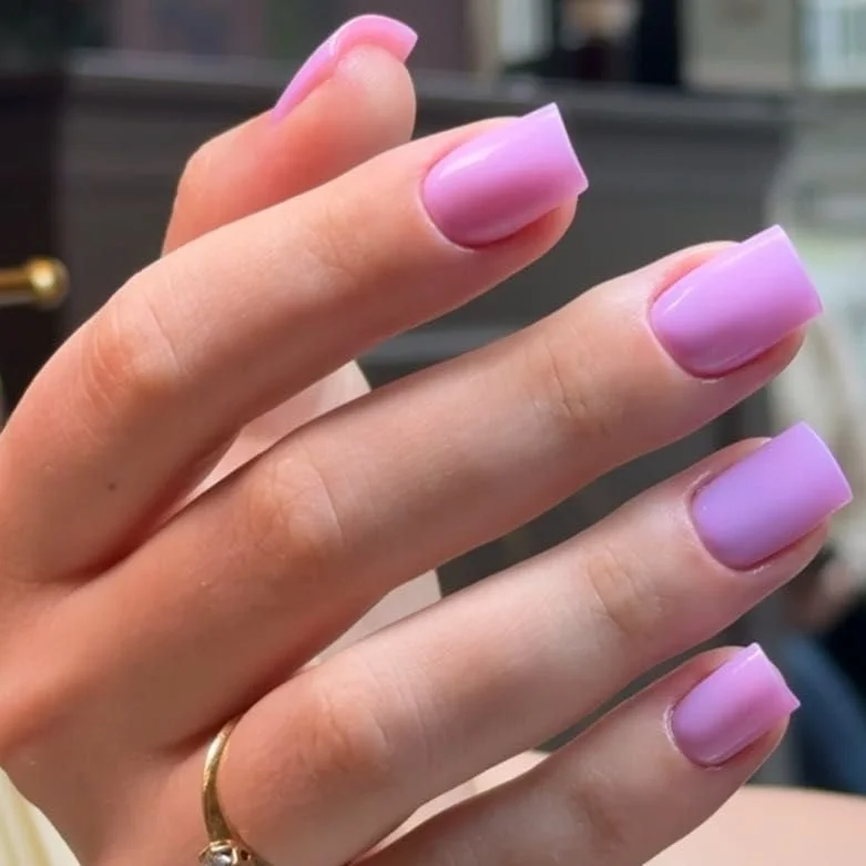 A close-up of a person's hand with neatly manicured nails painted in a soft lavender color, wearing a gold ring with a small gemstone.