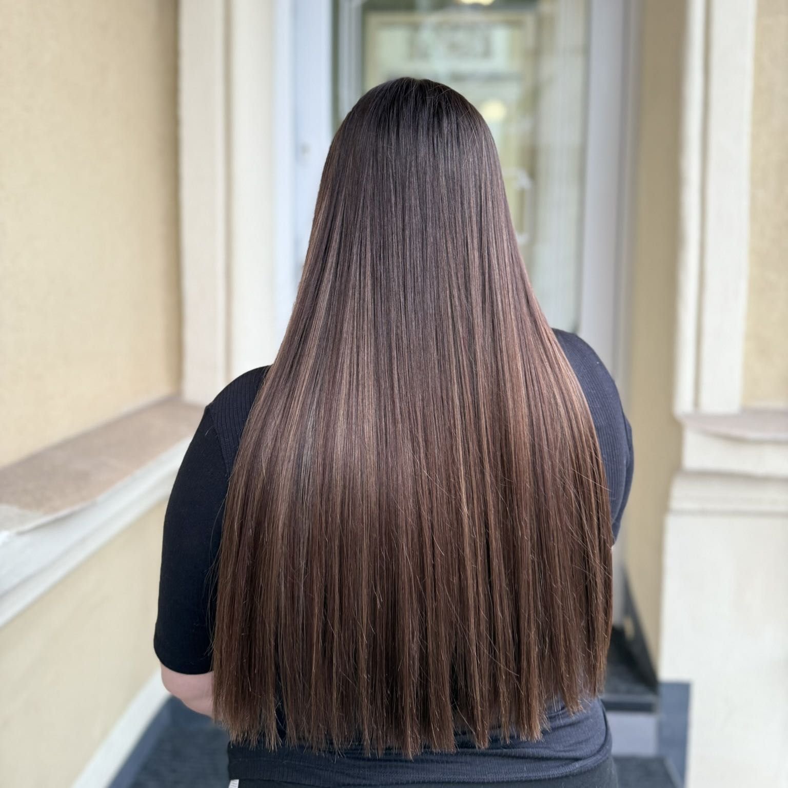 Back view of a woman with long, straight, silky brown hair standing indoors near a window.
