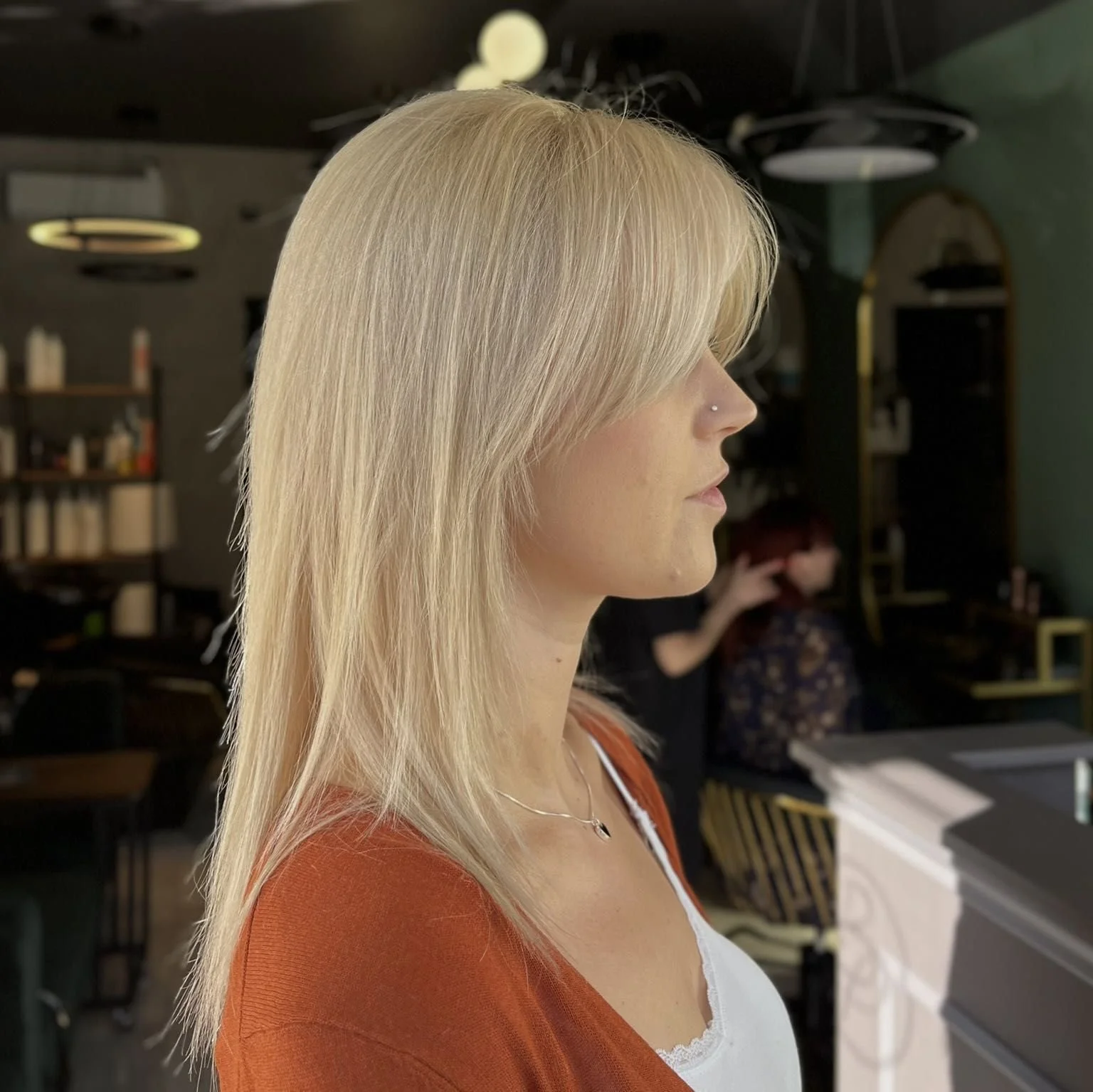 Side profile of a woman with blonde hair and a nose piercing, wearing a white top and an orange cardigan, standing indoors.