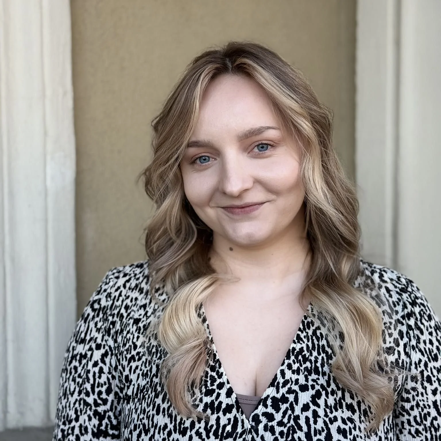 A young woman with shoulder-length, wavy blonde hair and blue eyes, smiling at the camera, standing outdoors in front of a beige wall with white trim.