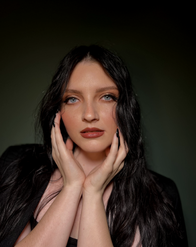 A woman with long black hair and blue eyes posing with her hands gently on her face against a dark background.