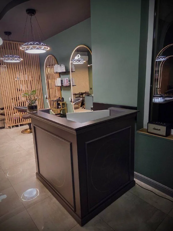 Modern reception desk with a white sink, dark wood paneling, and a black faucet, in a contemporary interior space with green walls, round mirrors, pendant lights, and decorative plants.