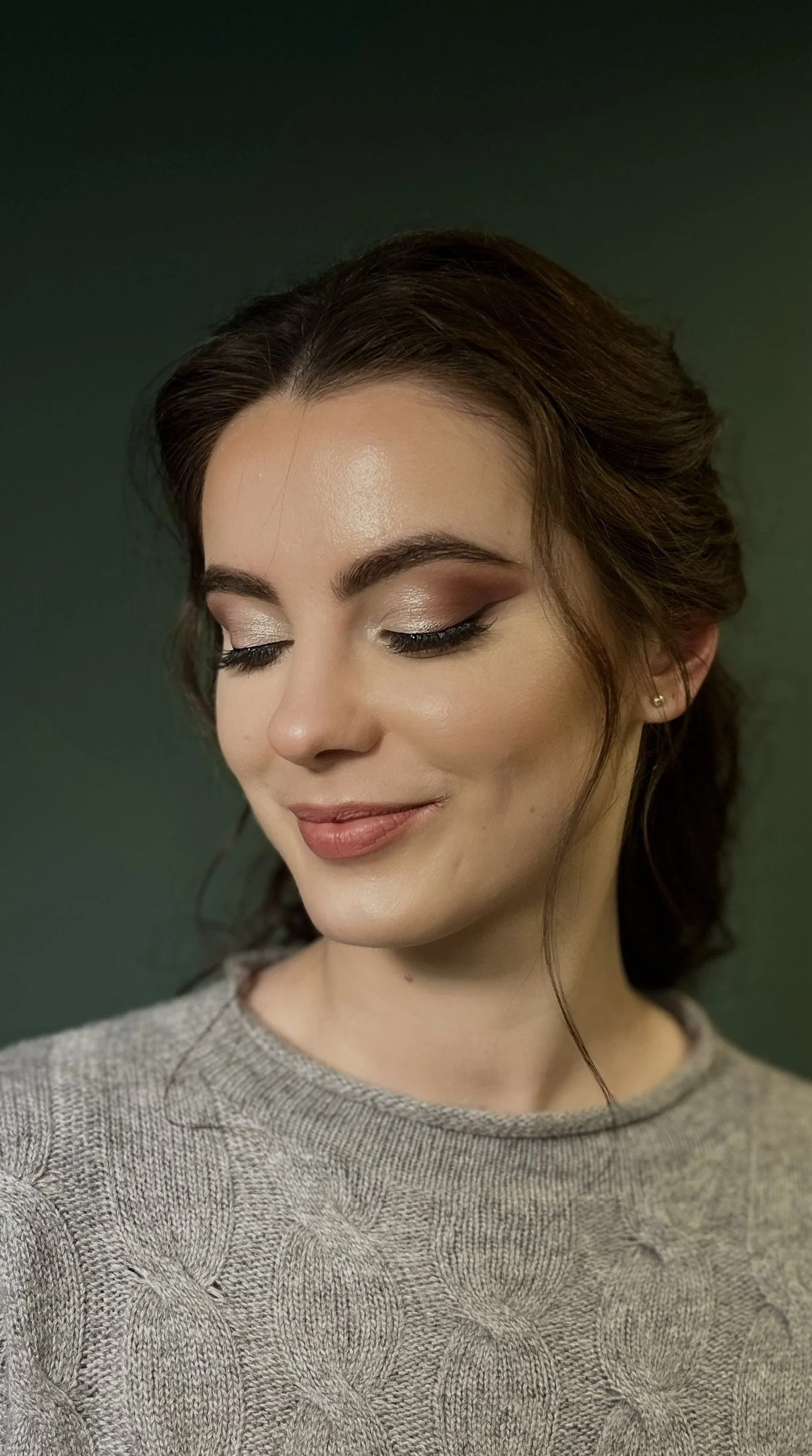 A woman with styled brown hair and makeup with neutral eyeshadow, winged eyeliner, and nude lipstick, wearing a gray knit sweater with a textured pattern, smiling softly against a green background.