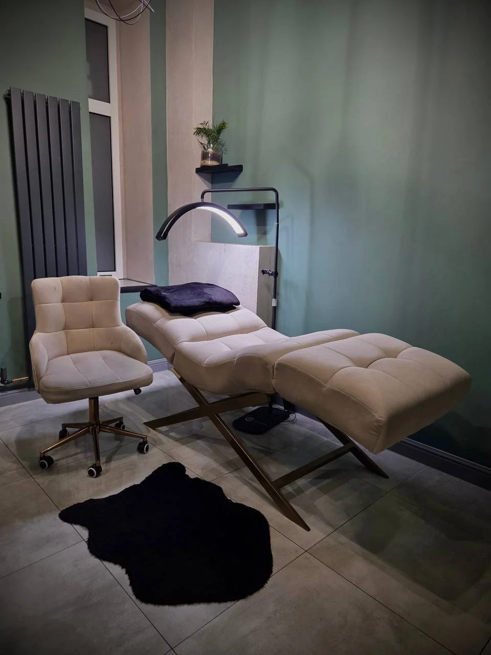 A modern room with a beige reclining chair and matching stool, a white office chair, a black night lamp, a small black rug, a potted plant on a wall shelf, and a gray wall with window and radiator.