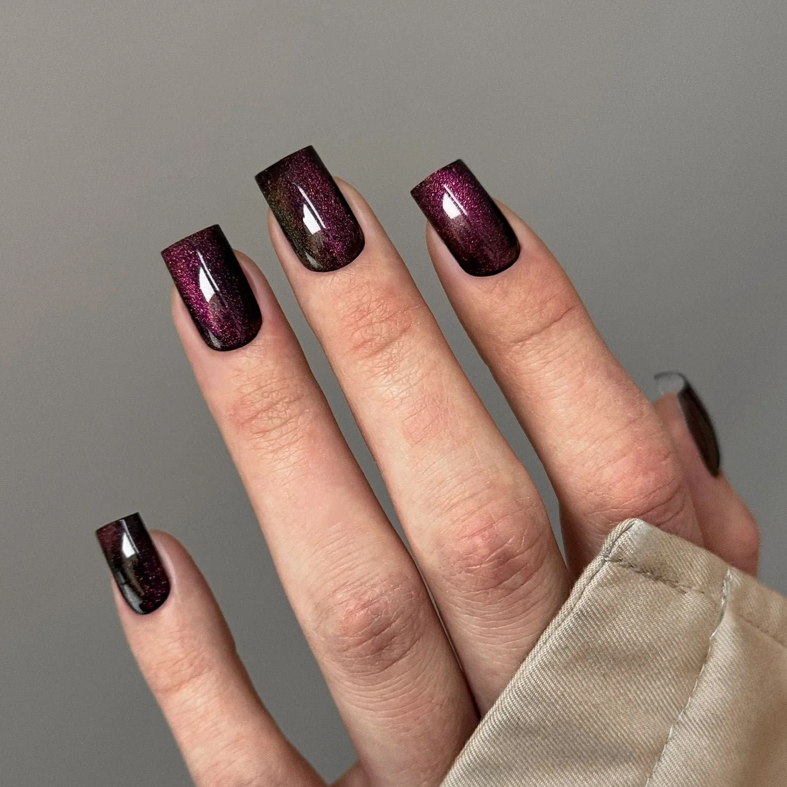 Close-up of a hand with five fingers, showing nails painted with dark purple to black nail polish with glitter, against a plain grey background.
