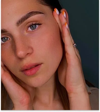 A young woman with clear skin and blue eyes, touching her face with both hands, wearing a ring on her finger and having orange and white nail polish.