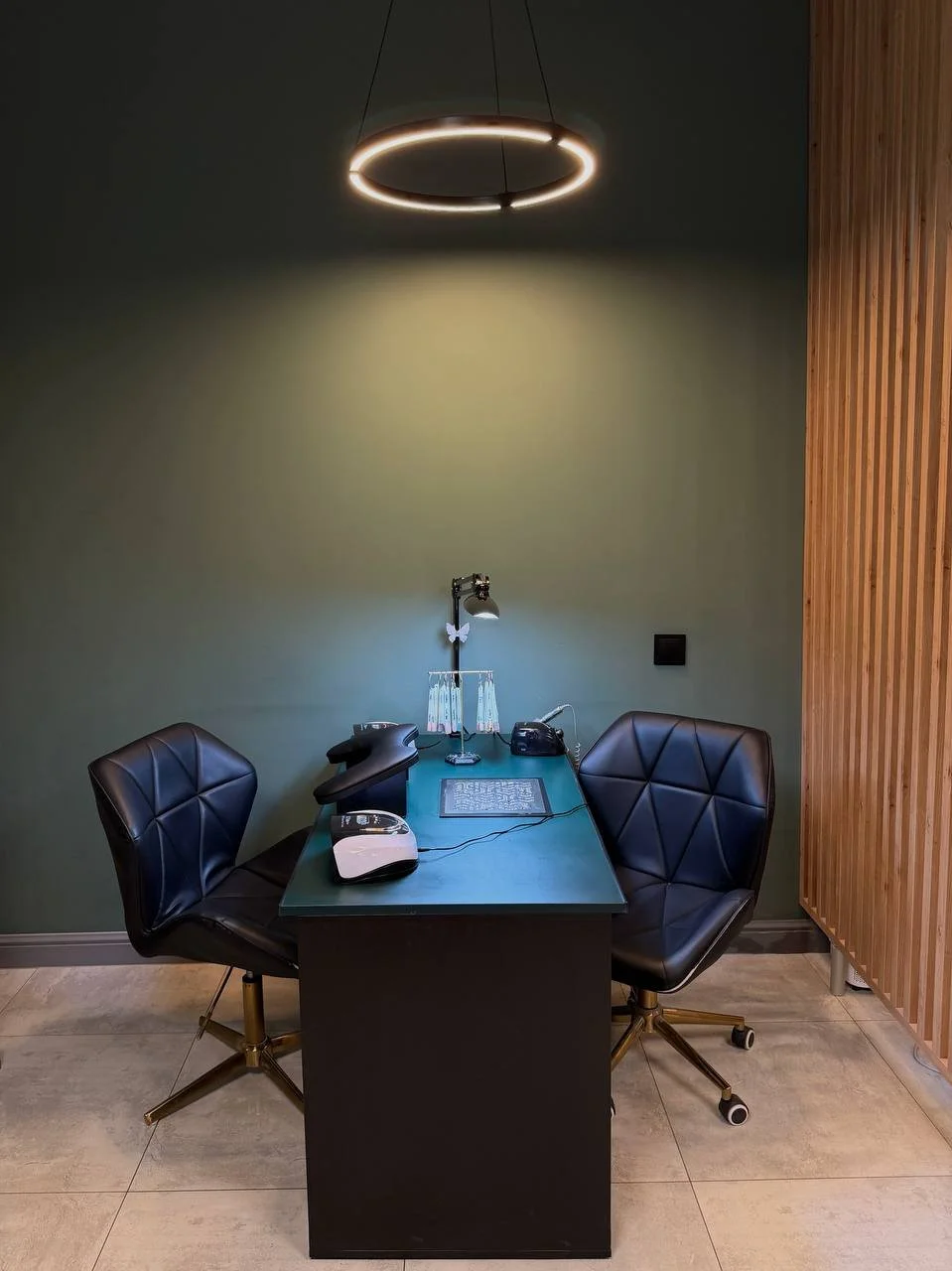 A modern office space with a black desk, two black chairs, a lamp, a telephone, and a few desktop items. A circular ceiling light fixture hangs overhead, and a wooden slat wall is visible on the right side.