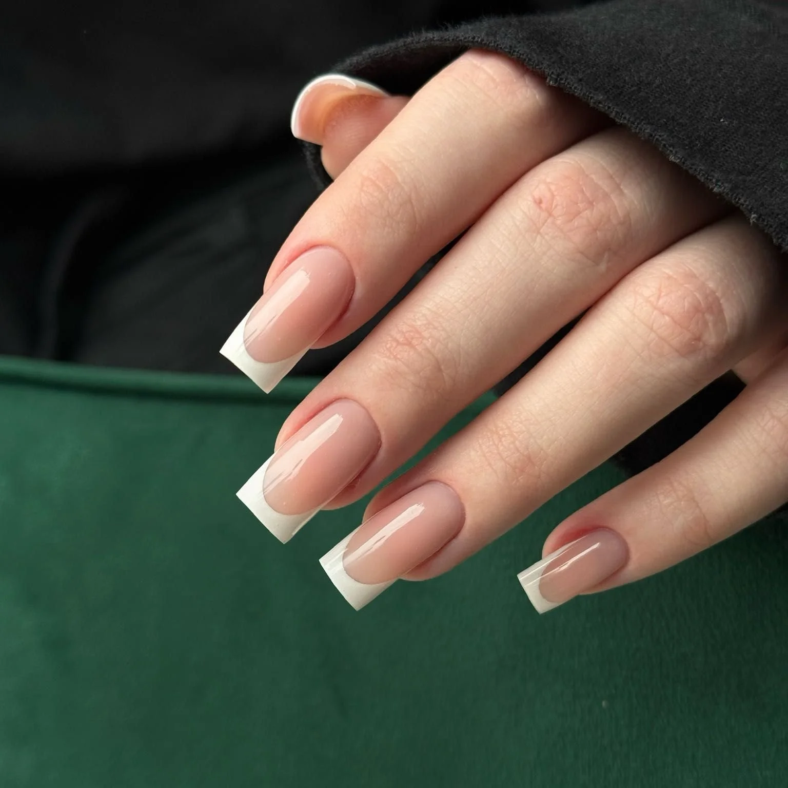 Close-up of a hand with natural-looking, acrylic French manicure nails, holding black fabric, against a green surface.