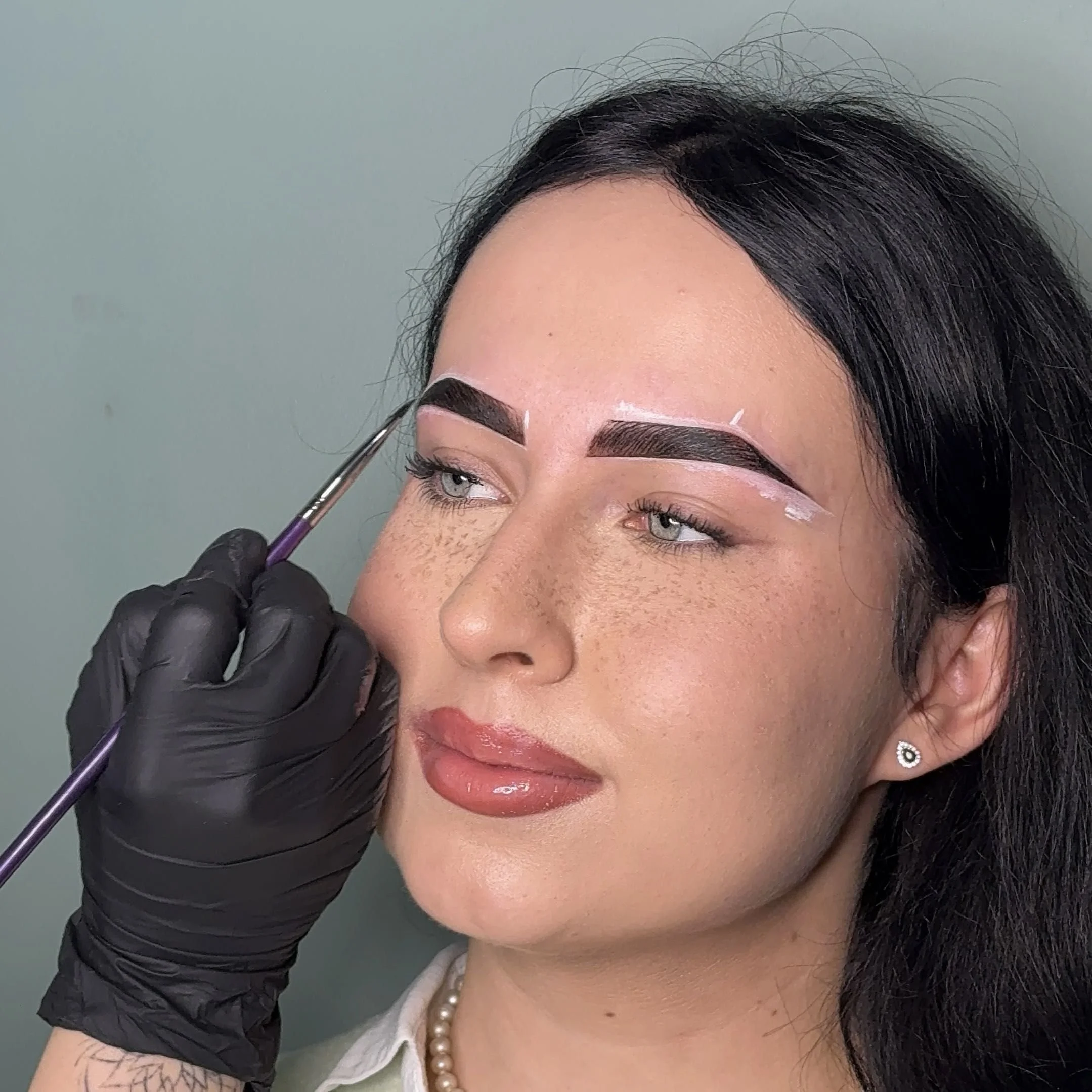 Woman with dark hair getting her eyebrows shaped or tattooed by a professional with black gloves and a fine brush.