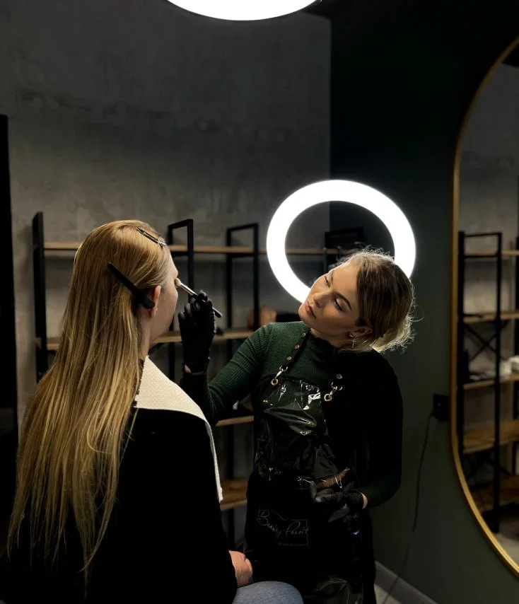 A woman with light brown hair receiving a makeup application from a makeup artist in a modern salon or studio, with shelves and a large mirror with ring light in the background.