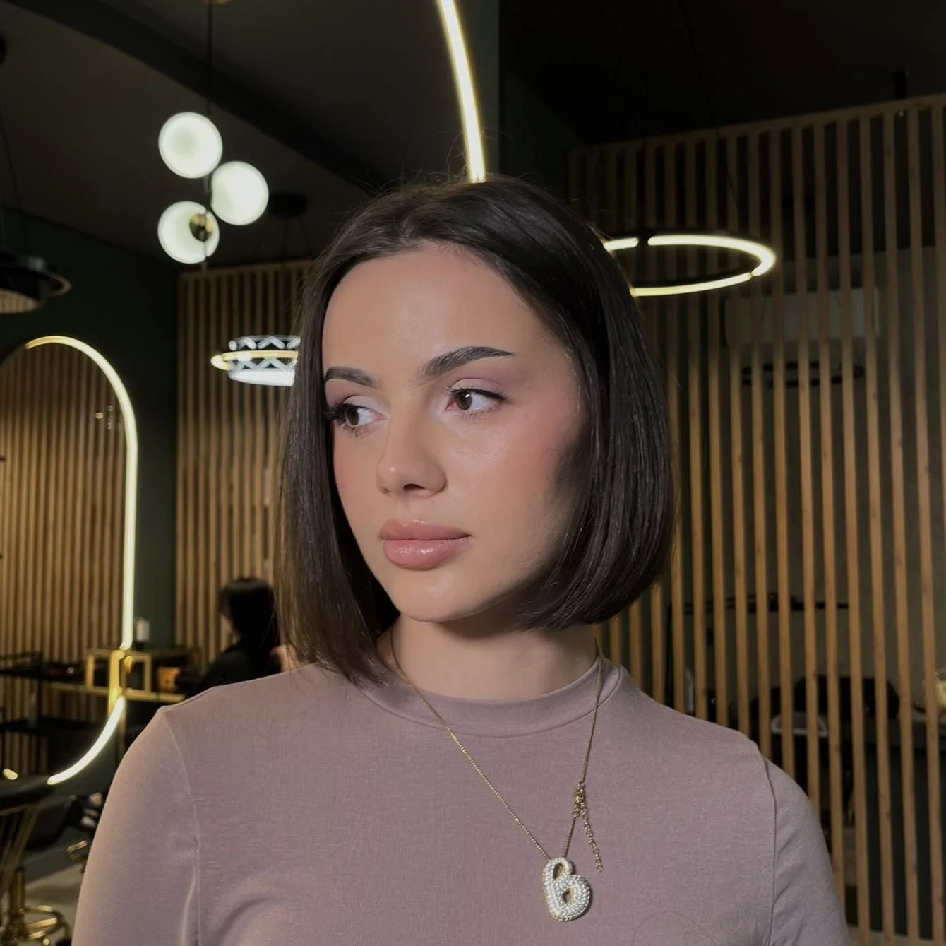 A woman with shoulder-length dark hair in a salon or modern interior, wearing a beige top and a gold necklace with a pendant shaped like the number six.