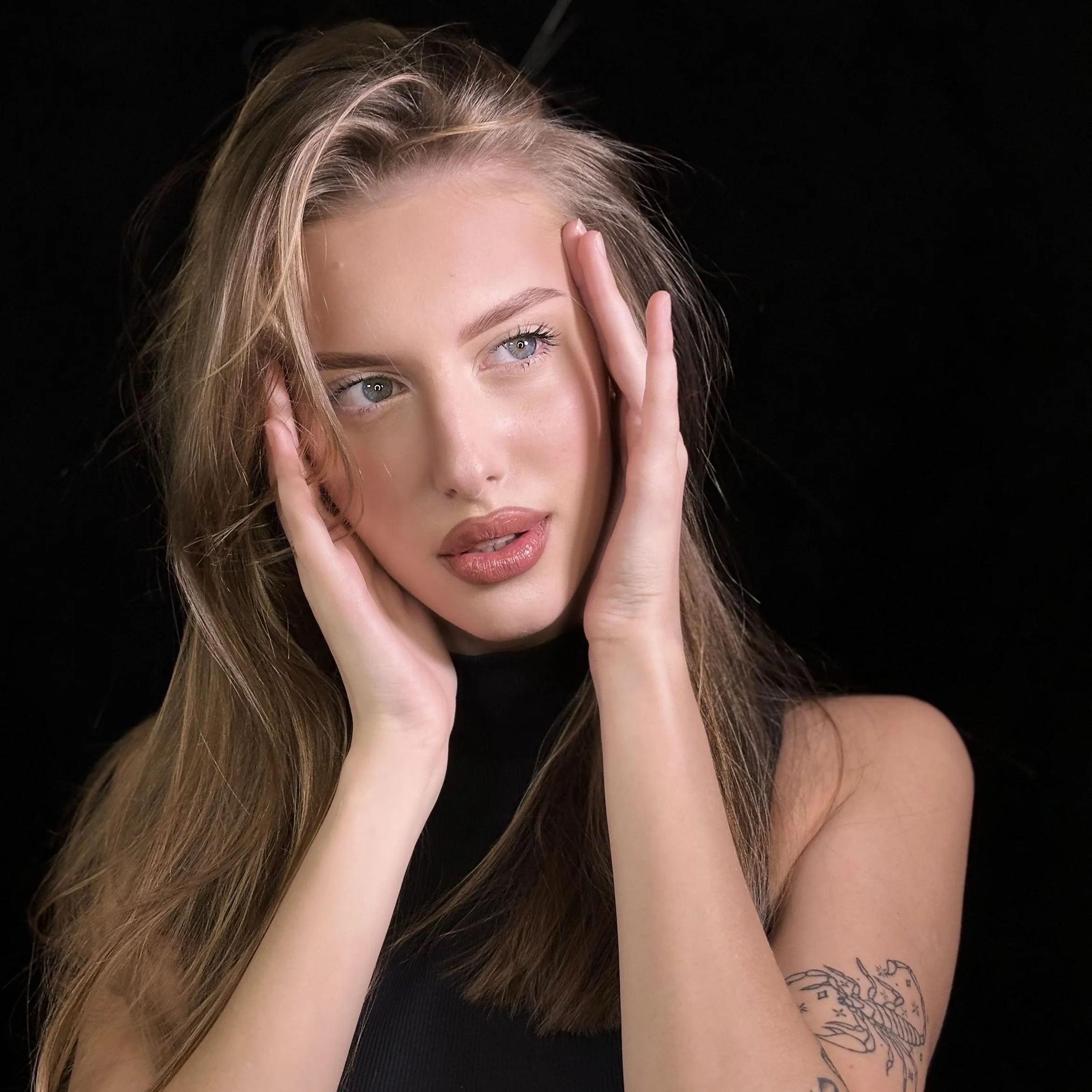 Close-up portrait of a young woman with long hair, holding her face with both hands, wearing a black sleeveless top, with a tattoo on her right arm, set against a black background.