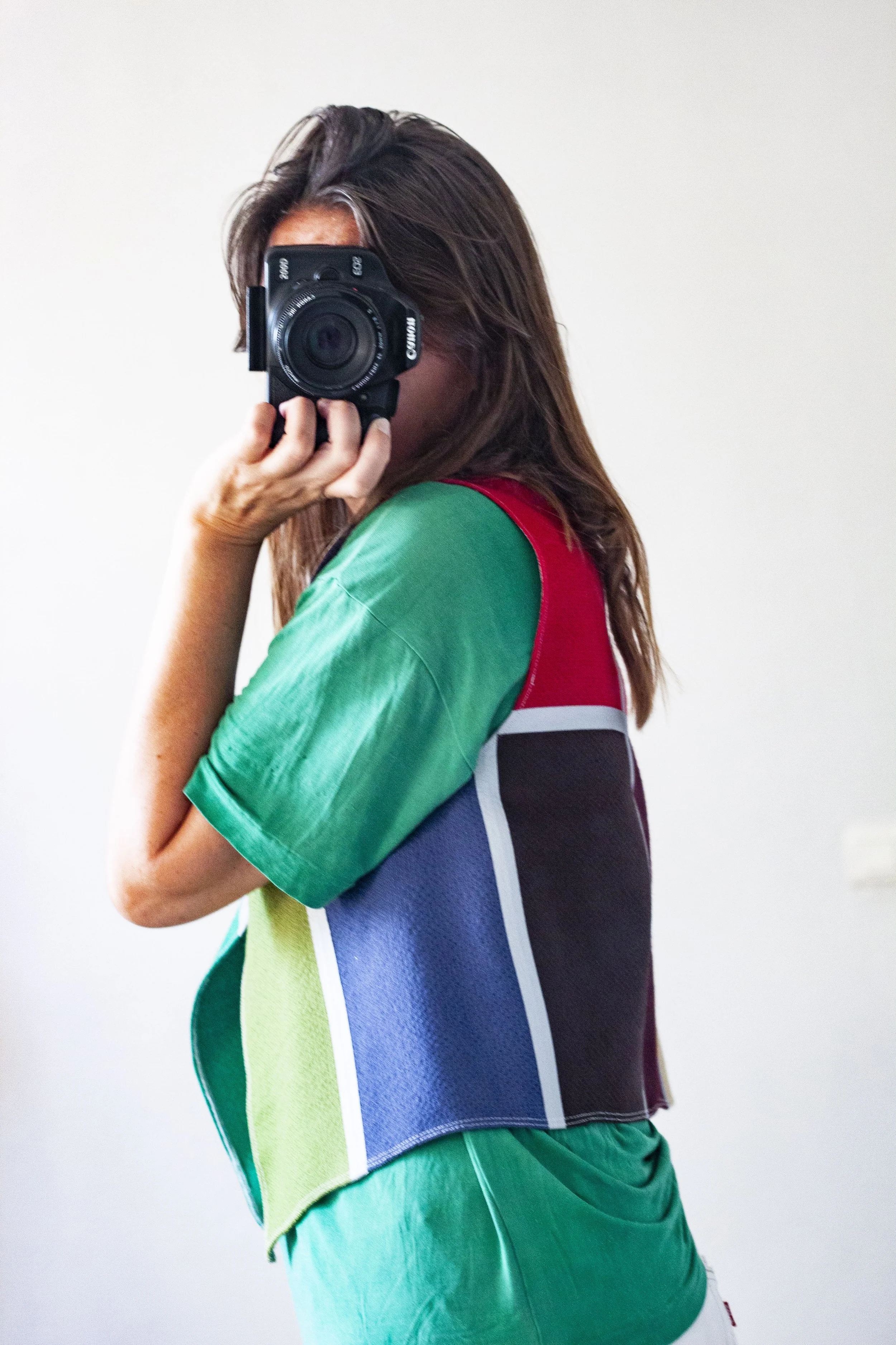 A woman with long brown hair wearing a multicolored shirt taking a selfie with a camera in a plain room.