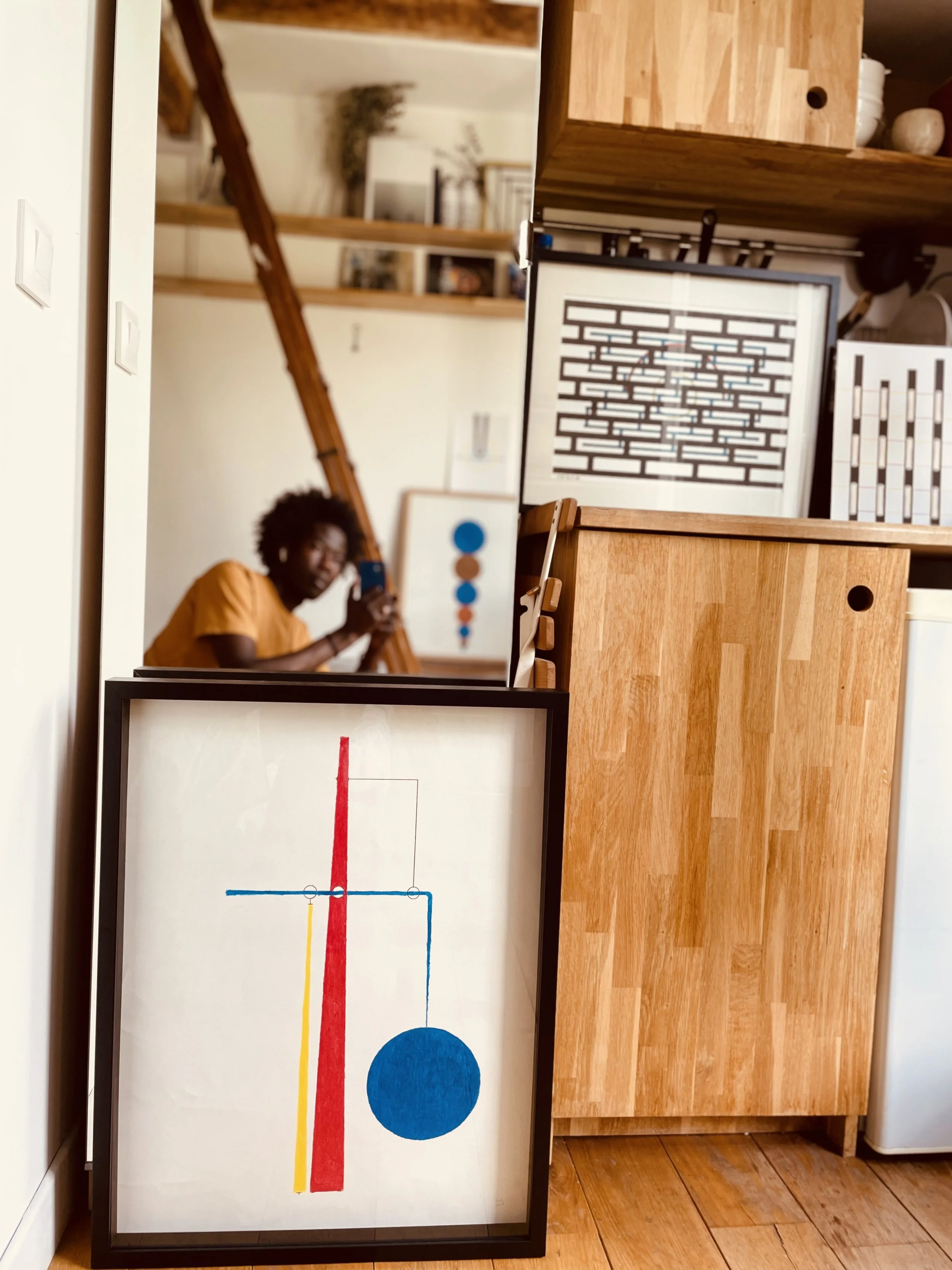 Part of a room featuring a framed abstract artwork with vertical red, yellow, and blue lines and a large blue circle, a mirror reflecting a person taking a photo with a smartphone, and wooden shelves and cabinetry.