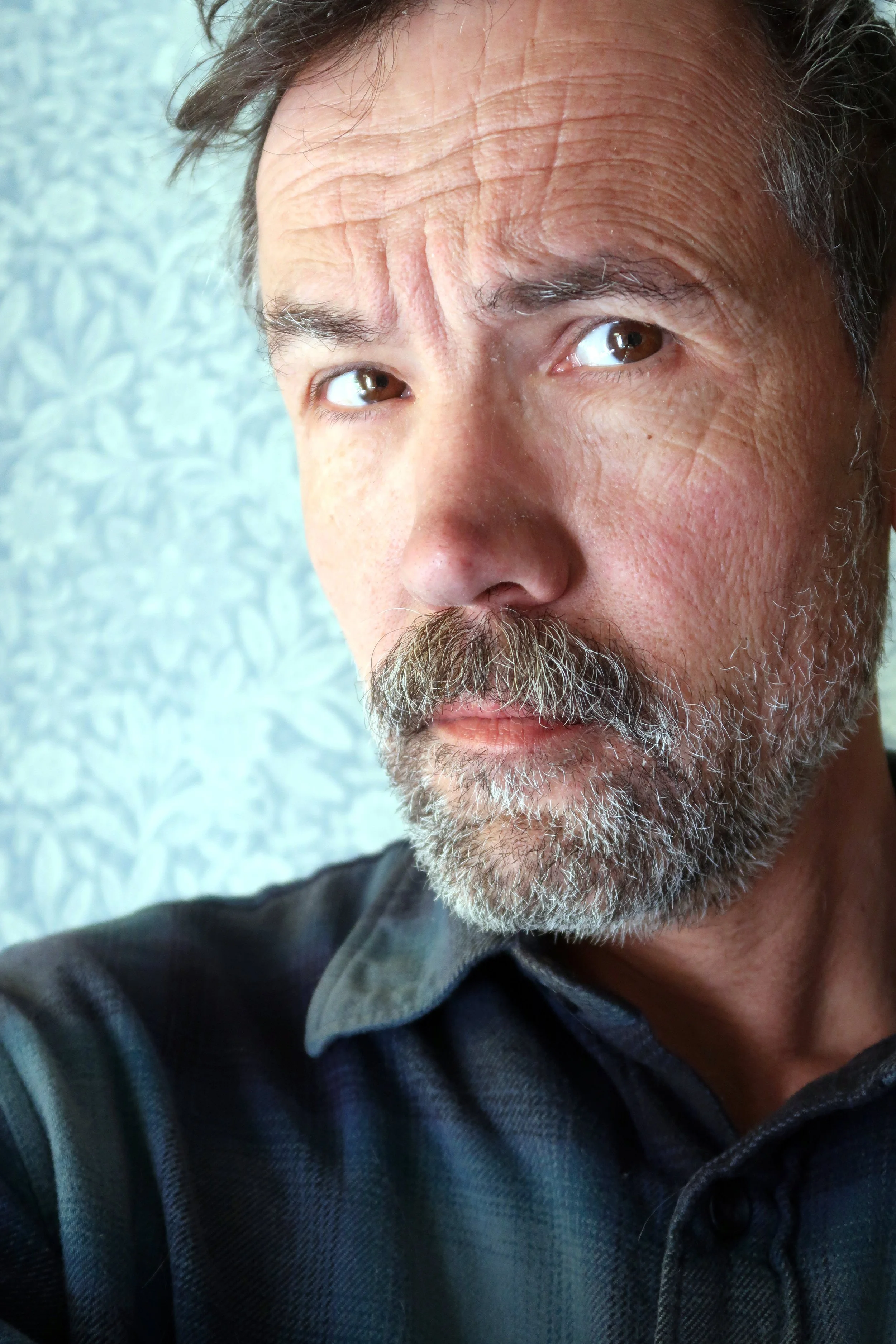 Close-up of a middle-aged man's face with a beard, showing wrinkles on his forehead and around his eyes. He has dark hair with some gray, and is wearing a dark collared shirt. The background has a light-colored textured wall.