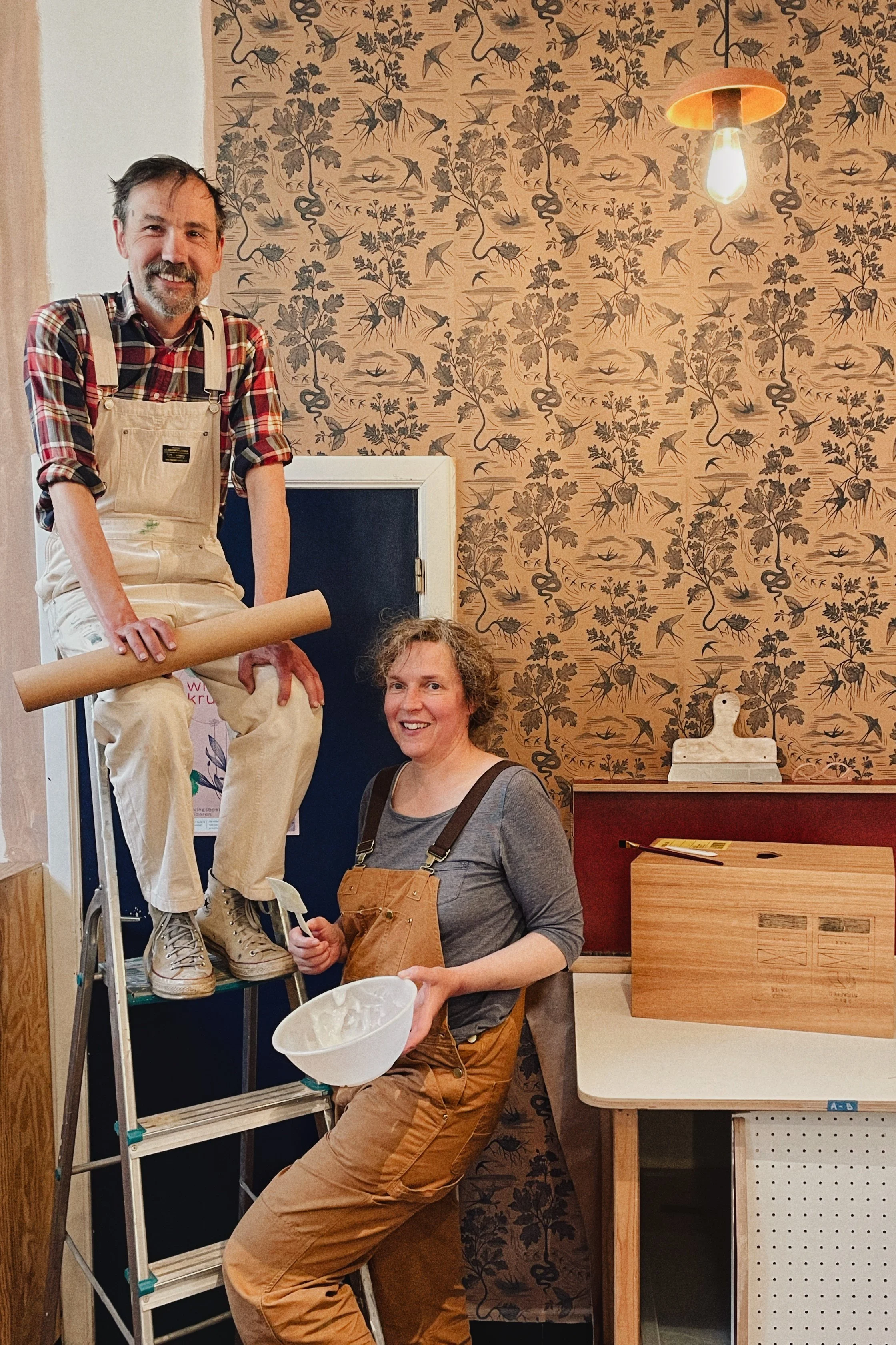 Two women in a room with patterned wallpaper, one standing on a ladder and the other sitting on a chair, holding a bowl and a spatula, during a crafting or decorating activity.