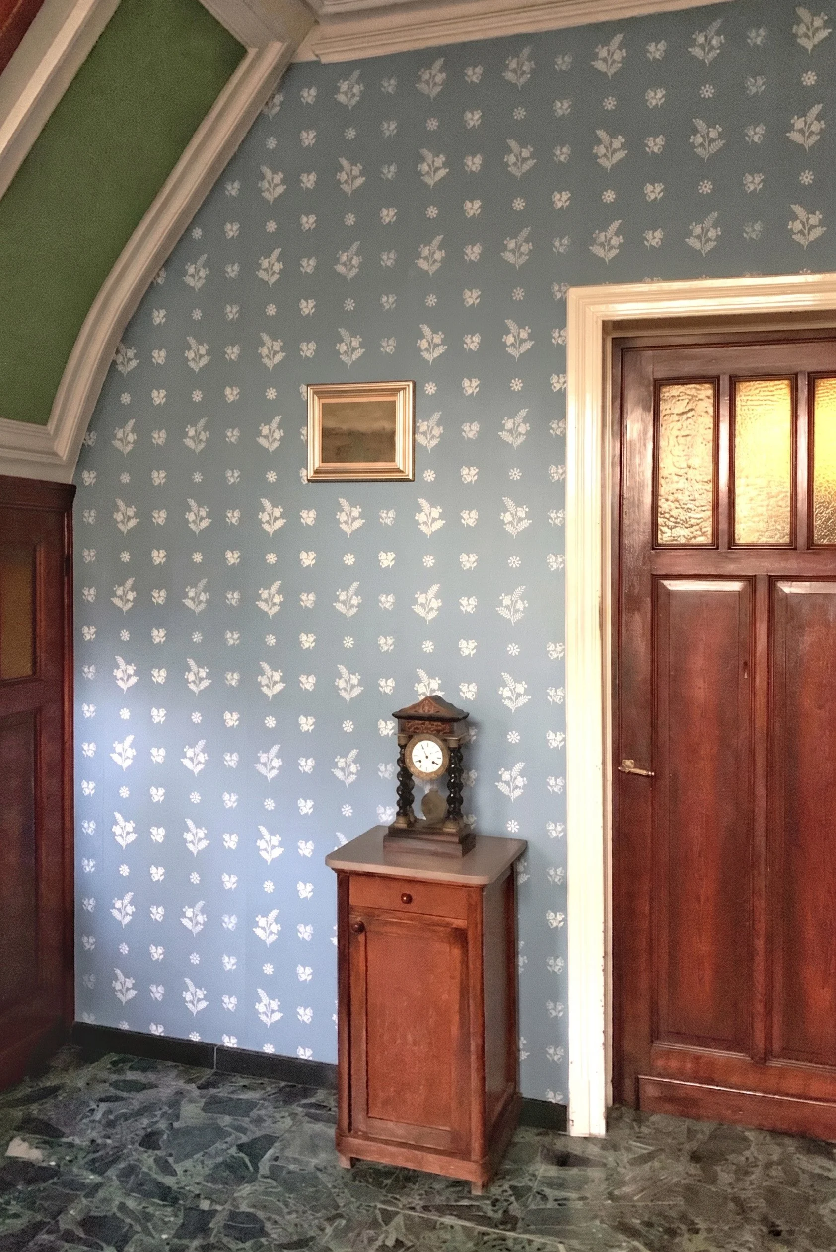 A room with a blue wallpapered wall decorated with white floral patterns, a small wooden table with a vintage clock, a framed picture, and wooden doors with textured glass panels, marble floor tiles.