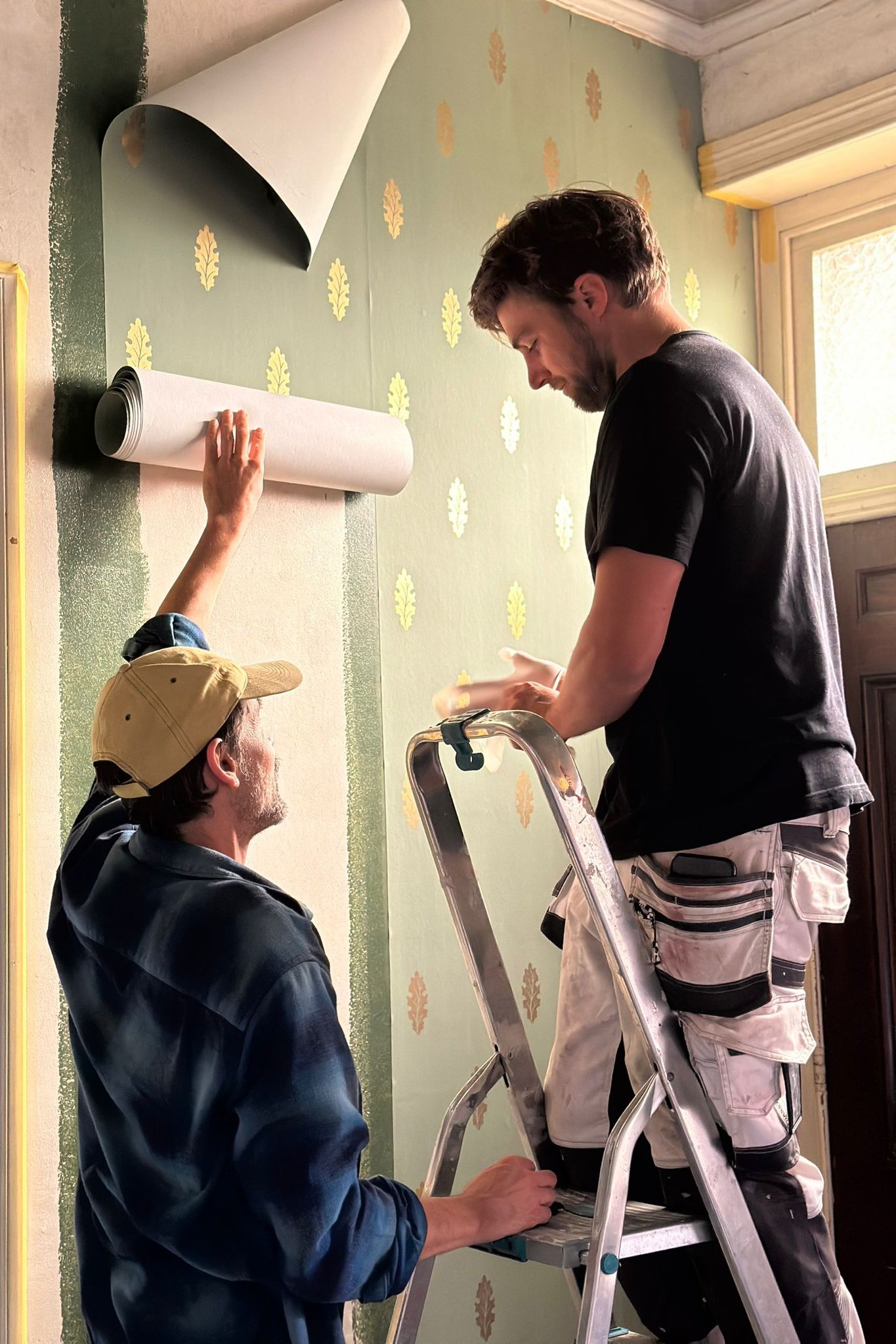 Two men, one older with a beard and one younger with dark hair, are working together on wallpaper installation. The older man is standing on a stepladder, holding a piece of wallpaper, while the younger man stands beside him, observing and assisting. A third person, wearing a beige baseball cap, is seated and holding the wallpaper roll. The background features a wall with green wallpaper decorated with yellow leaf patterns, and a window allowing natural light into the room.