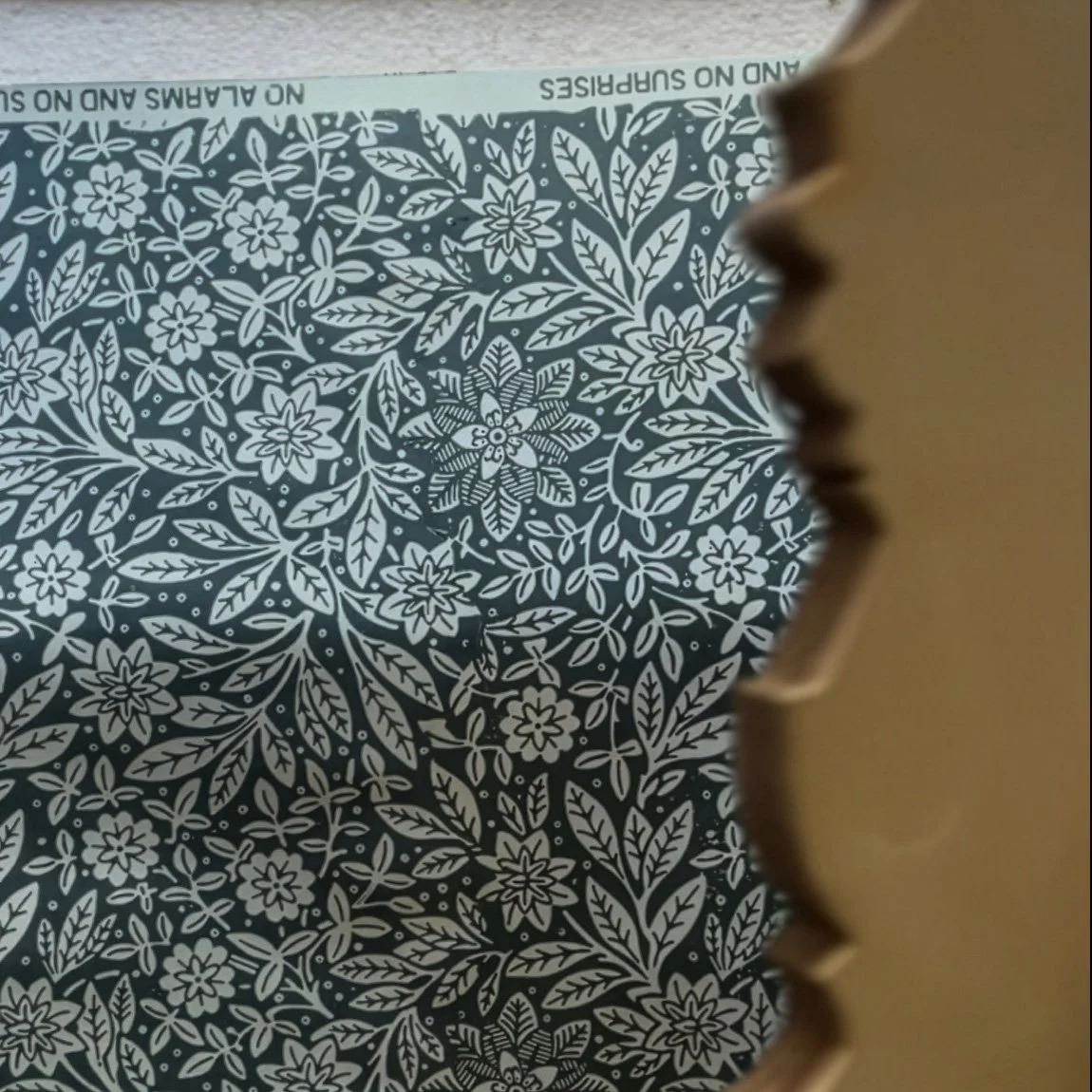 Close-up of a decorative cardboard packaging with white floral pattern, partially obscured by a brown scalloped-edged object in the foreground.