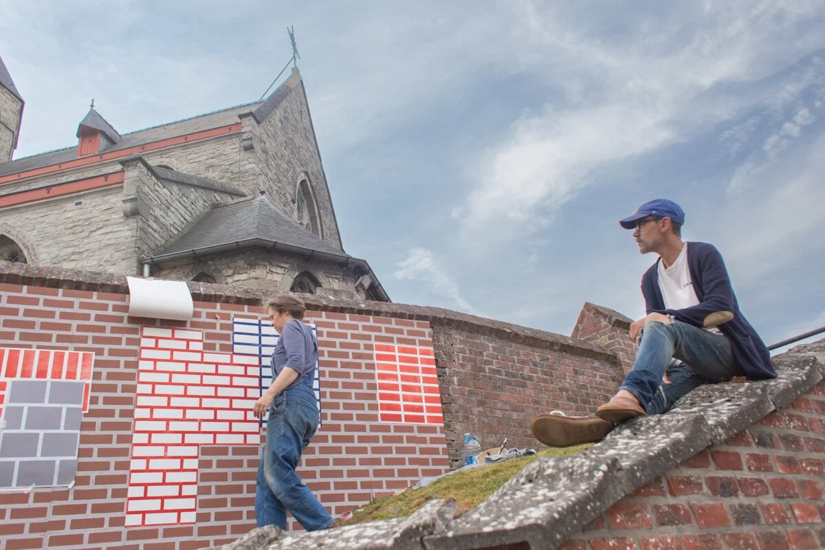 Two artists painting a mural of brick patterns on a brick wall outside a church building, with a man sitting on the roof watching.