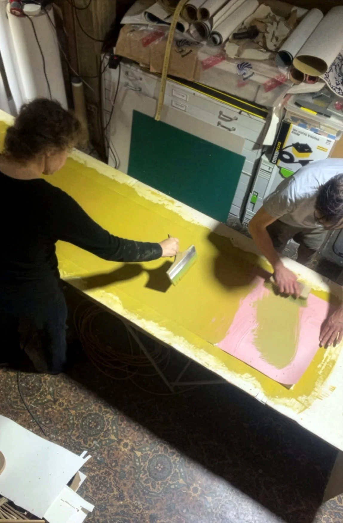 Two people working on a large yellow surface, painting a background for wallpaper, in a cluttered workshop with various supplies and rolled papers on the workbench.