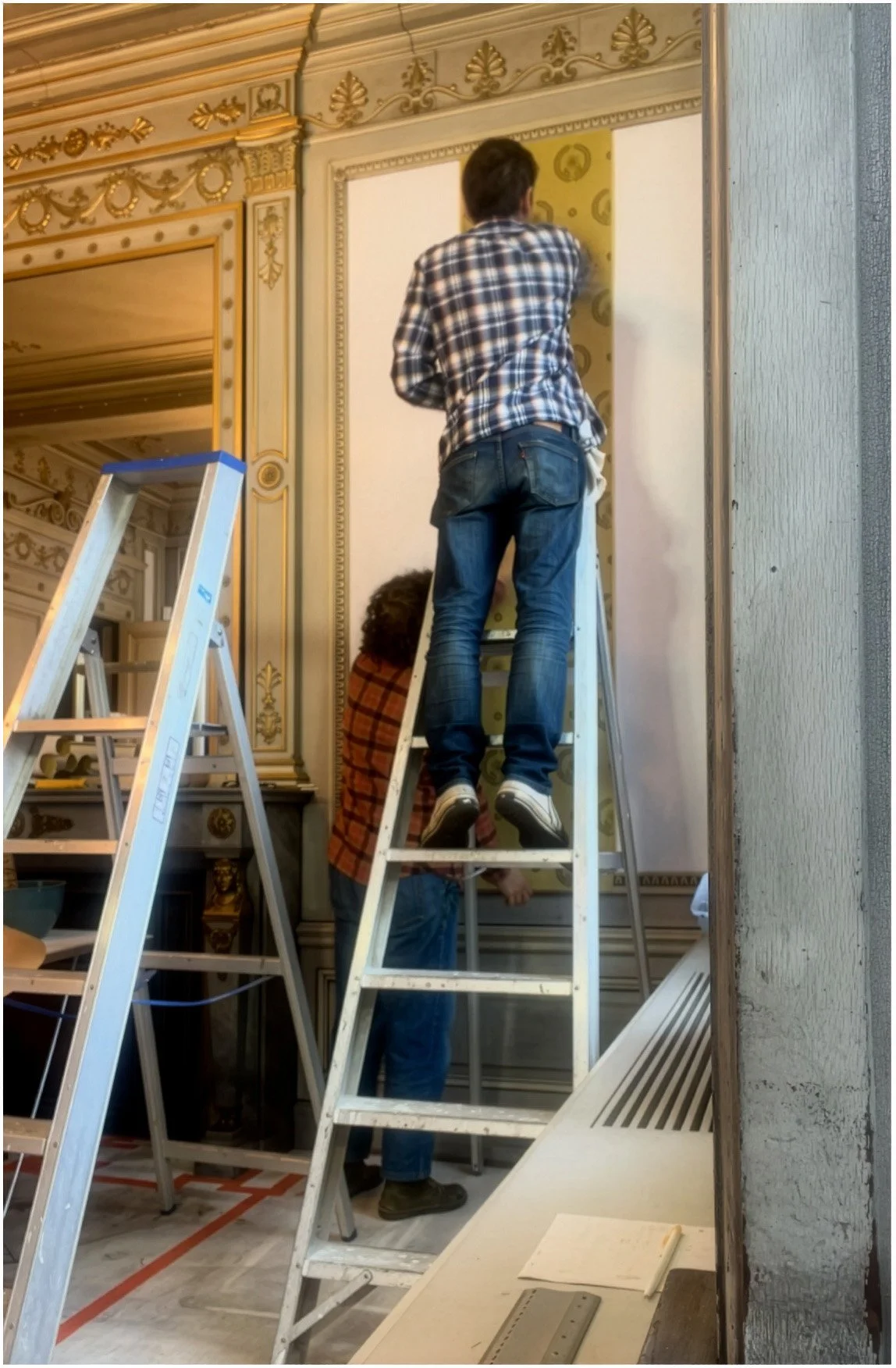 Two workers standing on ladders installing or repairing decorative wall paneling in an ornate, historic room with intricate moldings and gold accents.