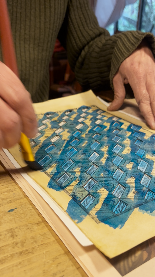 Person working on a detailed blue and yellow illustration of a map or diagram on paper, with hands and a pen visible.