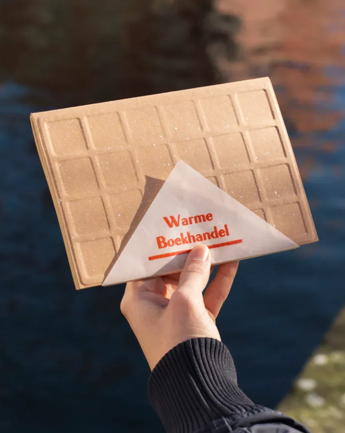 A person holding a chocolate bar with a paper wrapper that reads 'Warme Boekhandel' over a body of water.