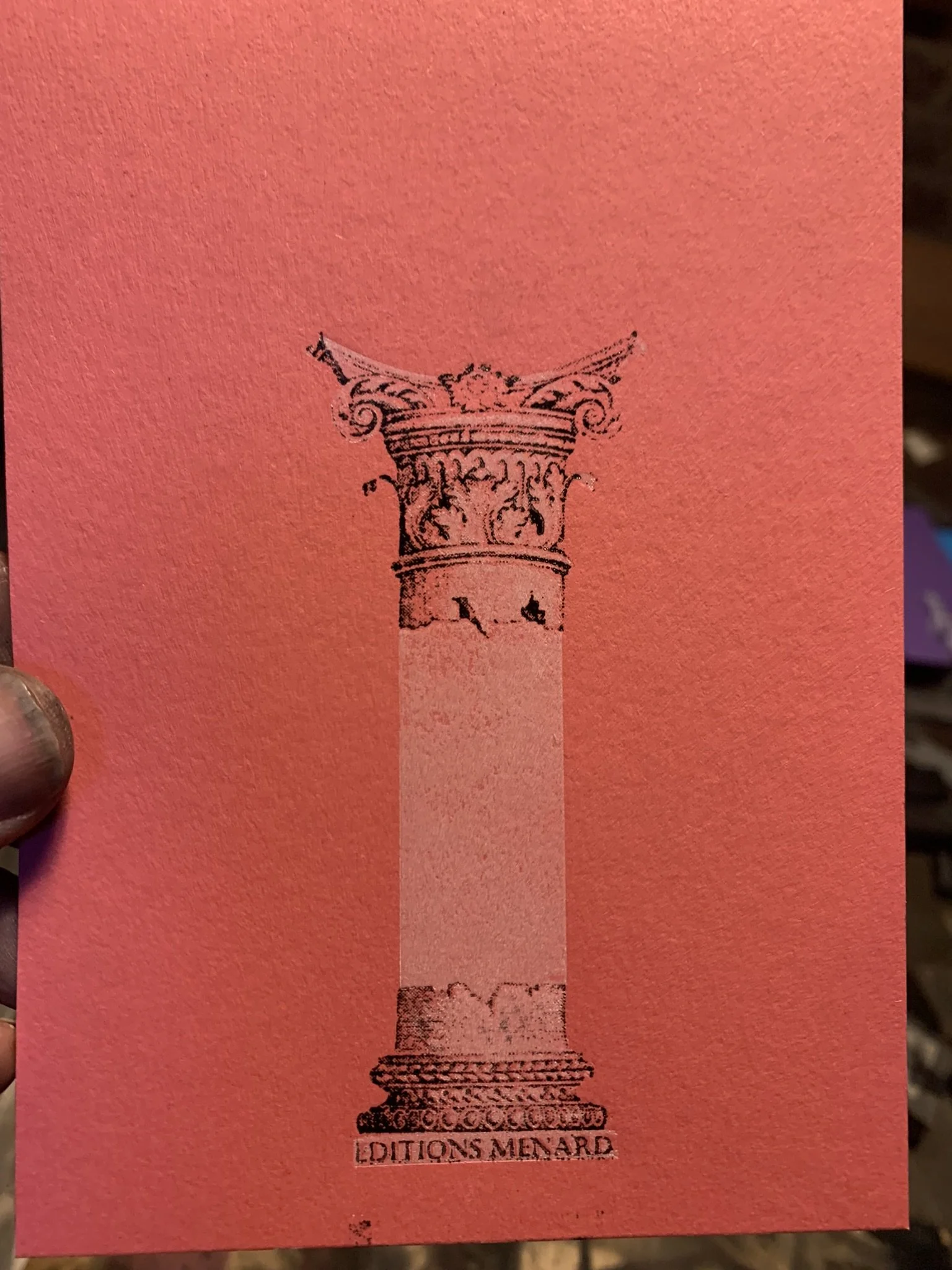 A red piece of paper with a black line drawing of a classical-style pedestal with three statues on it, and the text 'EDITION MENARD' underneath.