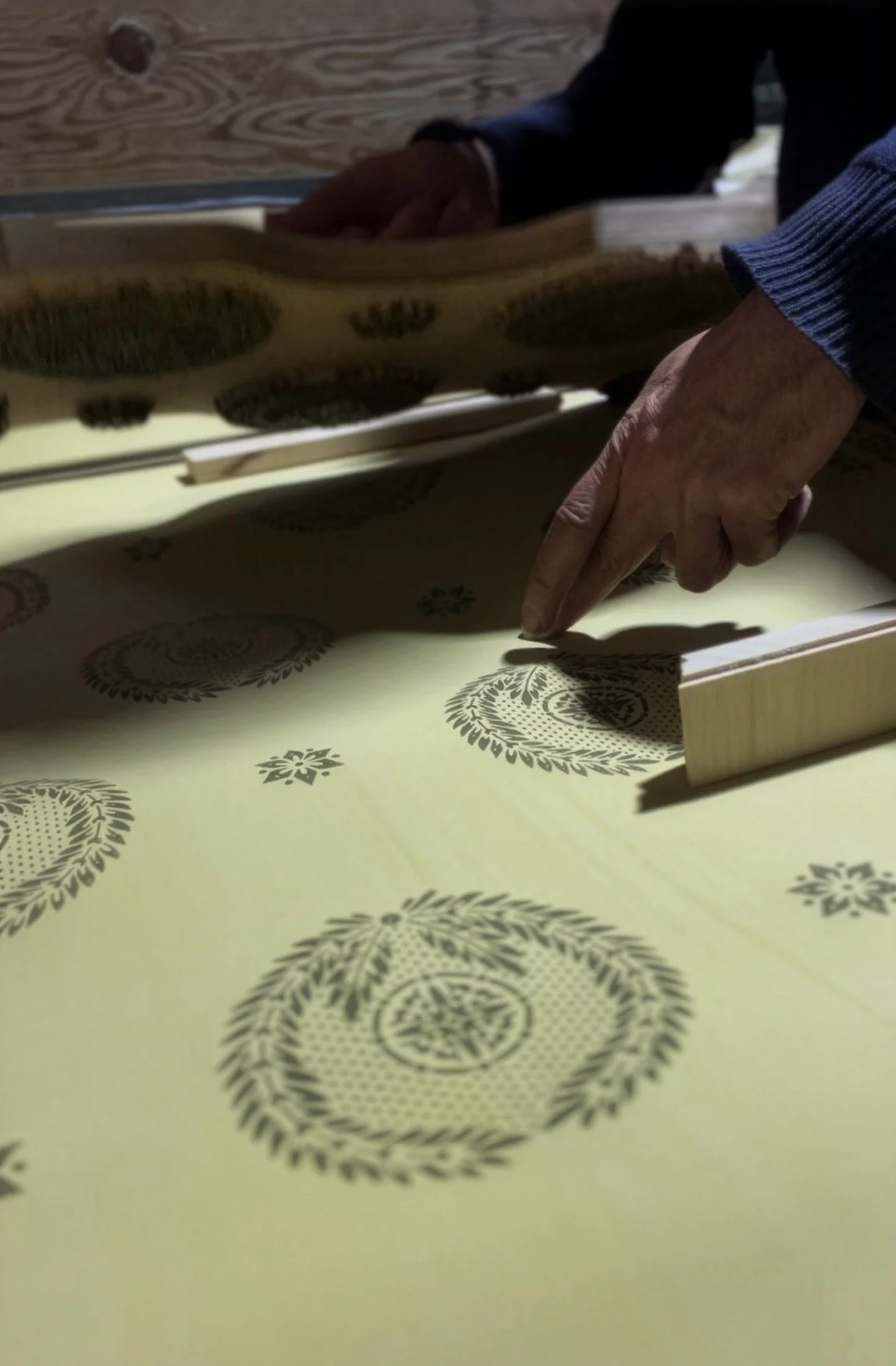 Close-up of hands pointing at a pattern on a piece of fabric or paper with a decorative design, possibly during a crafting or manufacturing process.