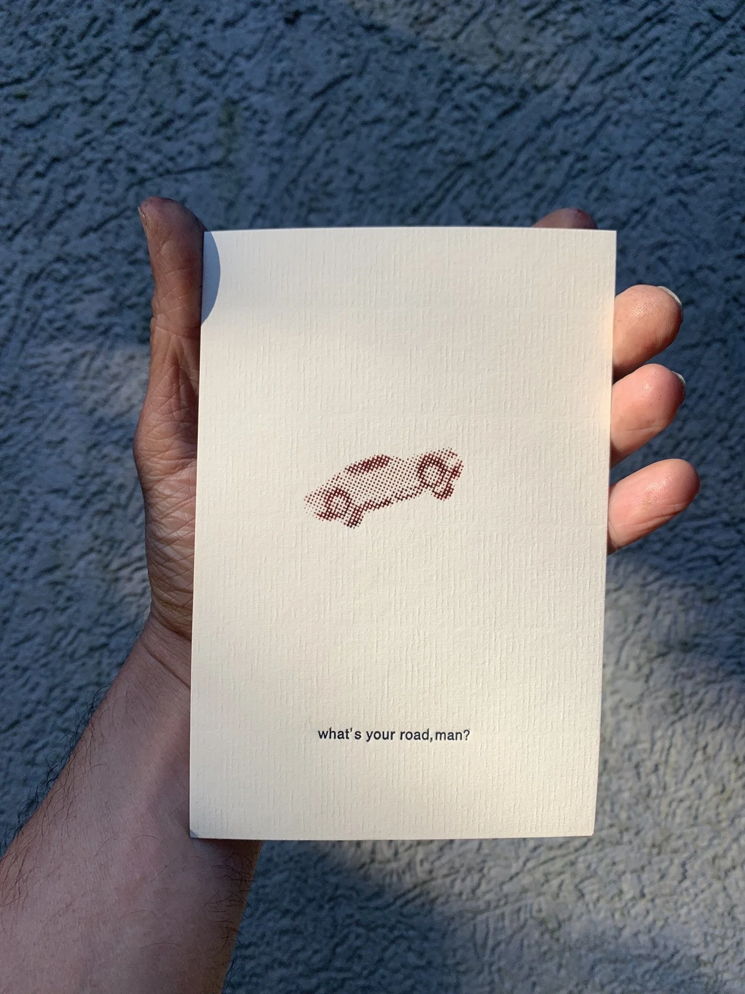 Holding a white card with an illustration of a red car and the text "what's your road, man?" printed at the bottom, against a textured dark blue background.