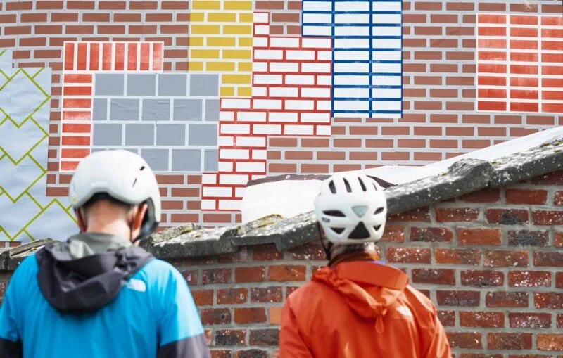 Two cyclists wearing helmets and jackets standing in front of a graffiti wall resembling a colorful brick and block pattern.