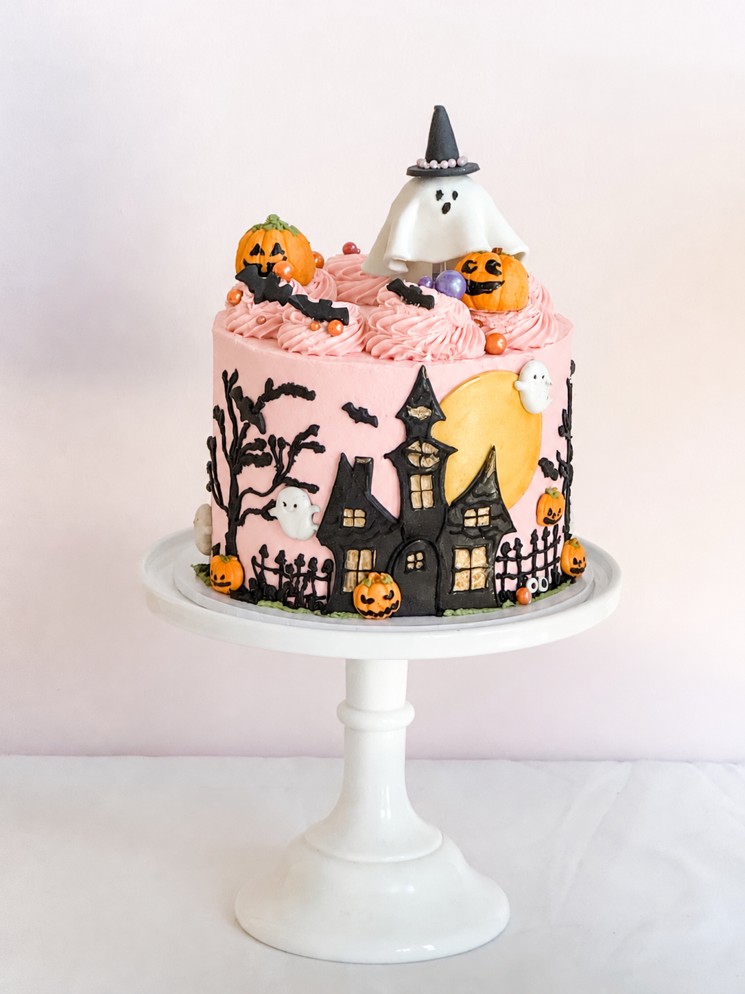 Pink Haunted Soirée Halloween cake by Sweet Stacy's Palm Beach featuring a black haunted mansion, golden moon, pumpkins, bats, and a ghost wearing a witch hat