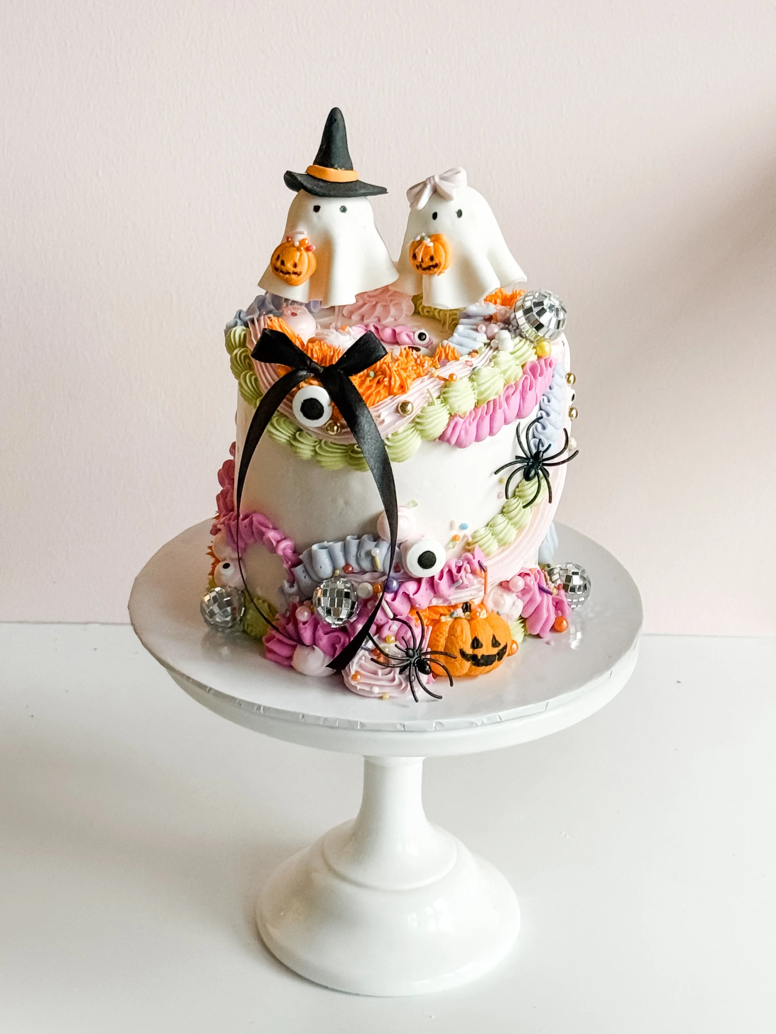 Boo's & Bows Halloween cake by Sweet Stacy's Palm Beach with two white ghost toppers, pastel buttercream piping, black bows, mini disco balls and pumpkins