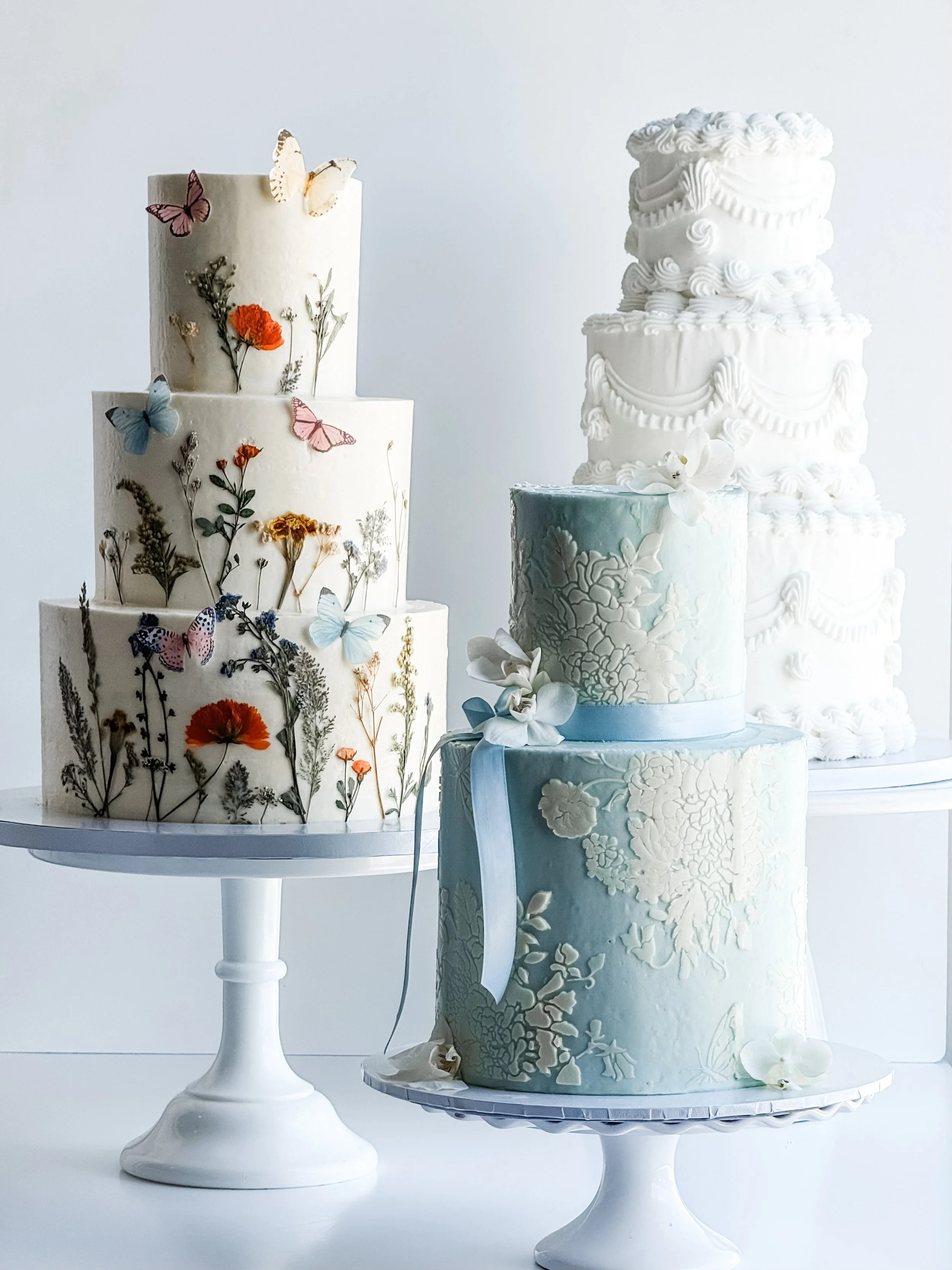 three tiered wedding cakes on cake stands - one decorated with butterflies, greenery and florals, one a light blue with white lace looking appliqués, and the last all white with vintage style frosting